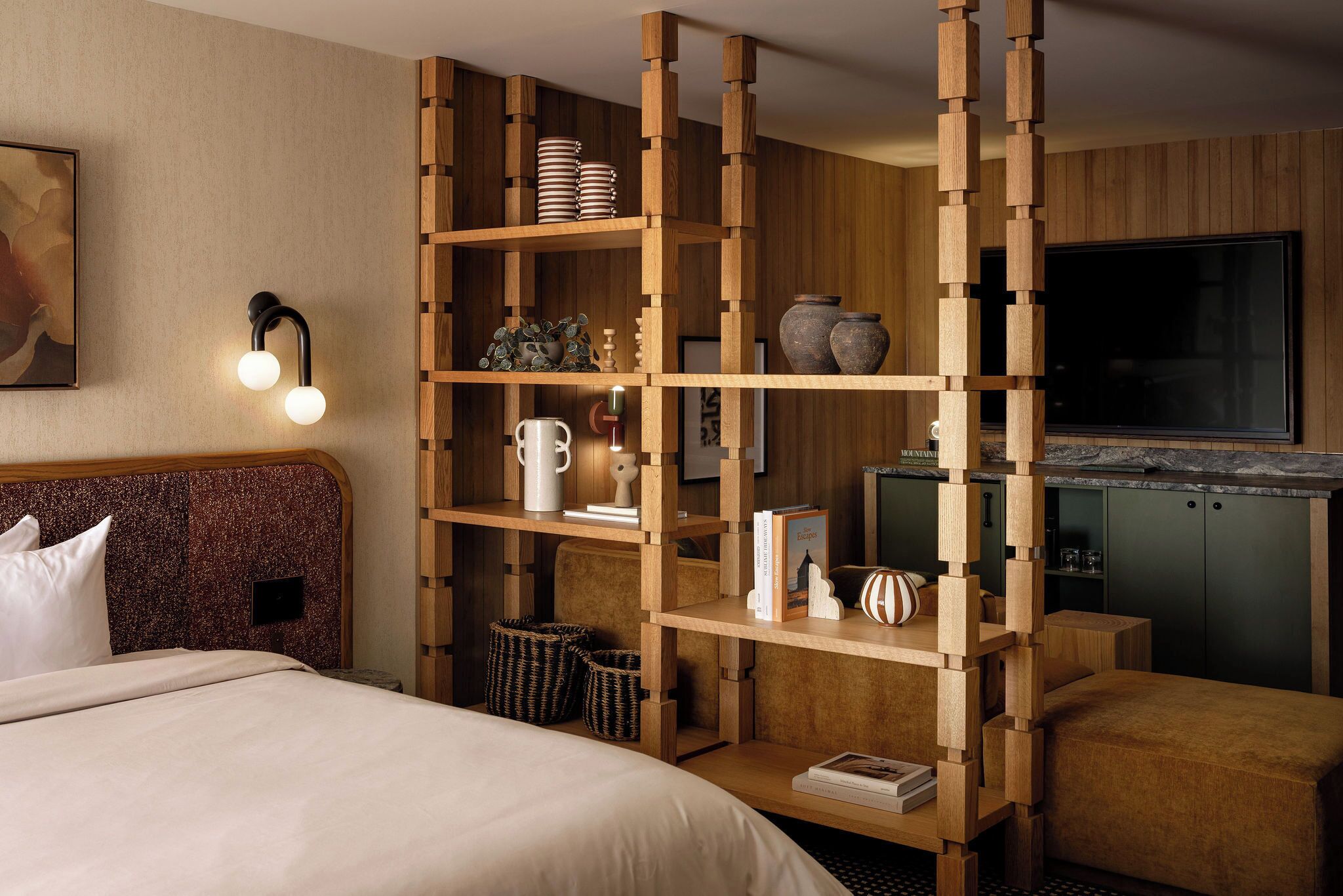 A hotel room with a wooden bookshelf, a bed, a TV, and a kitchen area.