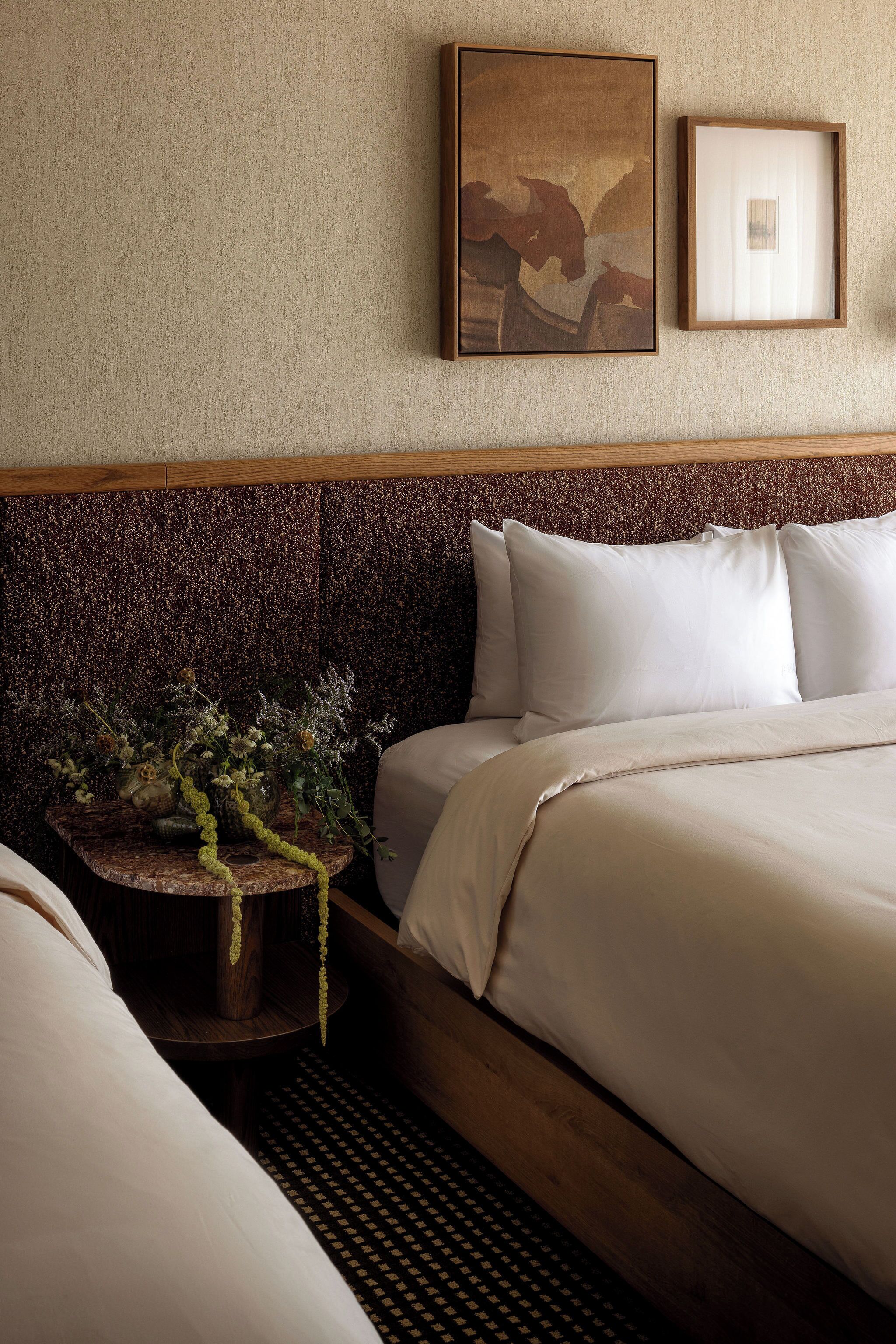 A hotel room with a bed, a headboard, a wall-mounted artwork, and a small table with a vase.