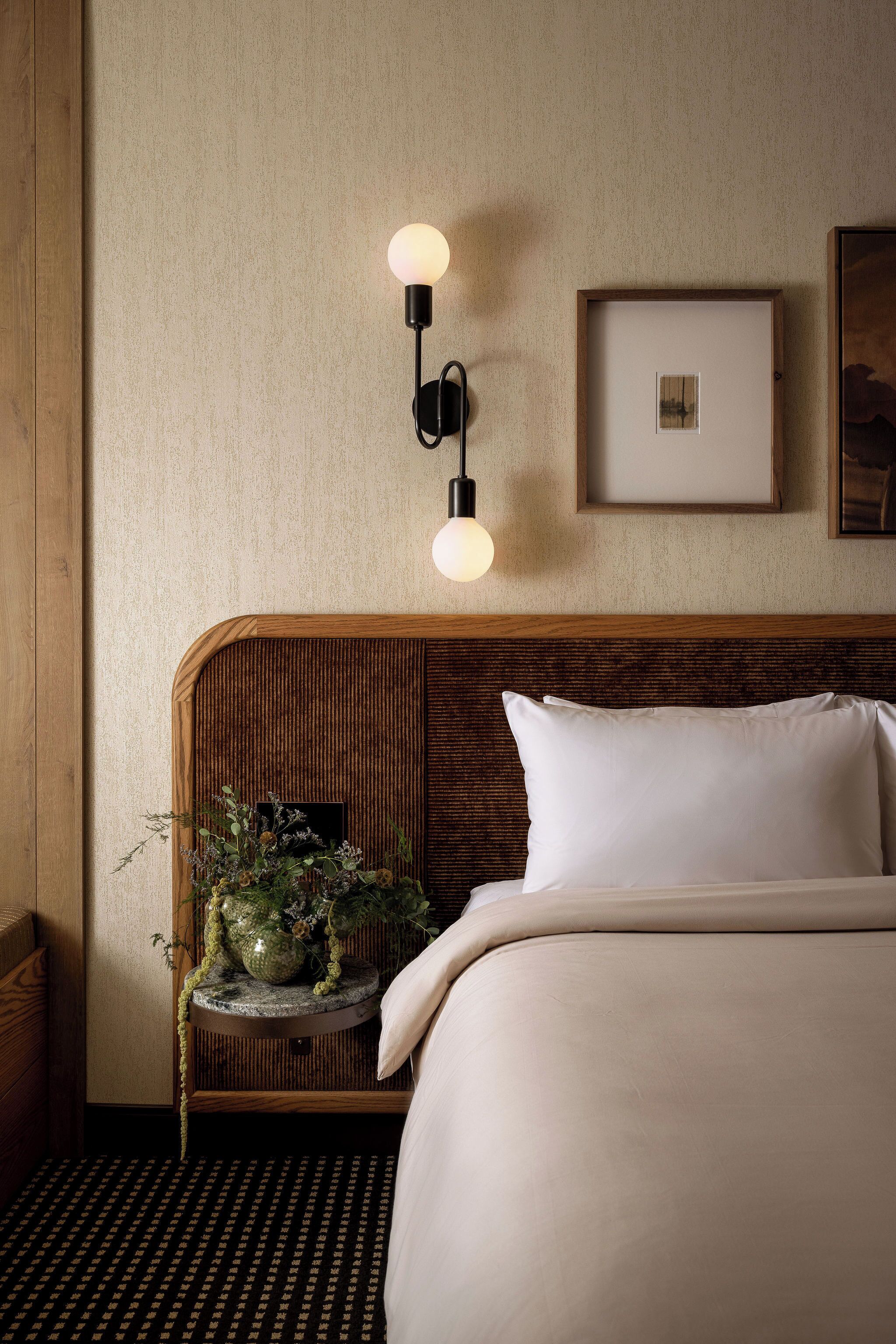 A hotel room with a wooden headboard, a round bedside table with a plant, a framed picture on the wall, and a bed with white bedding.