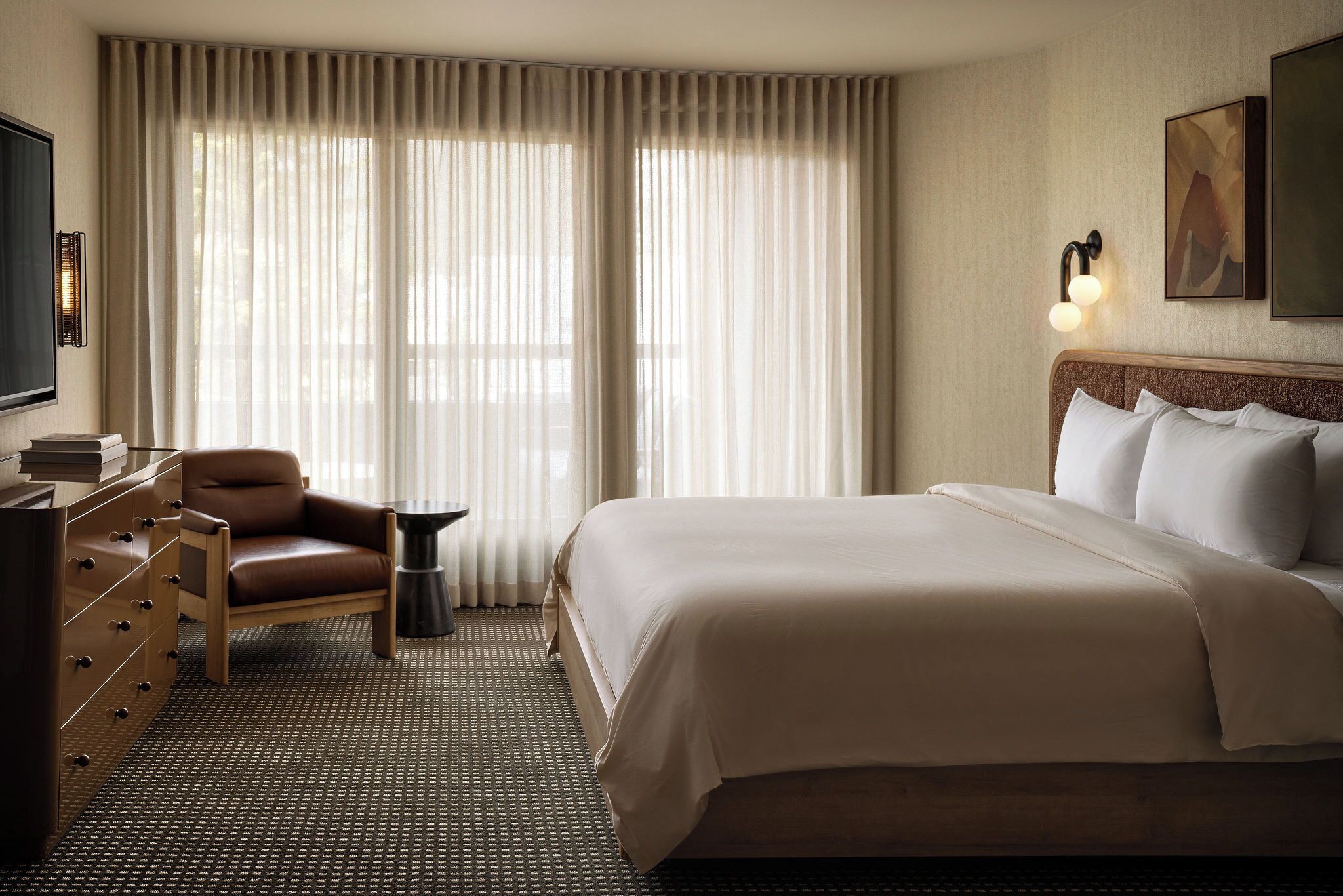A hotel room with a bed, a desk with a chair, a television, and a large window with curtains.