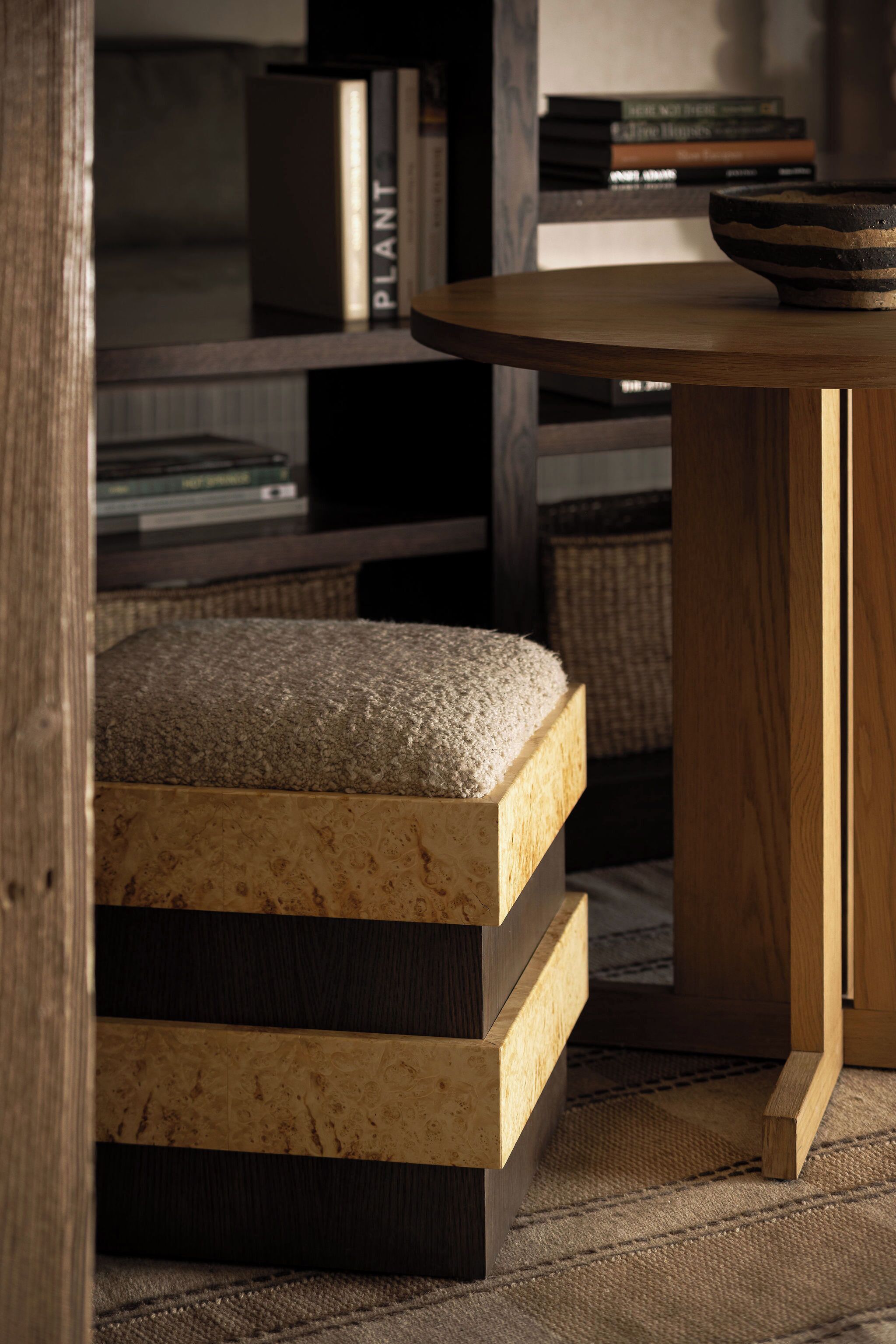 A wooden ottoman with a textured top and a lower shelf, a round wooden table, and a bookshelf with books and decorative items.