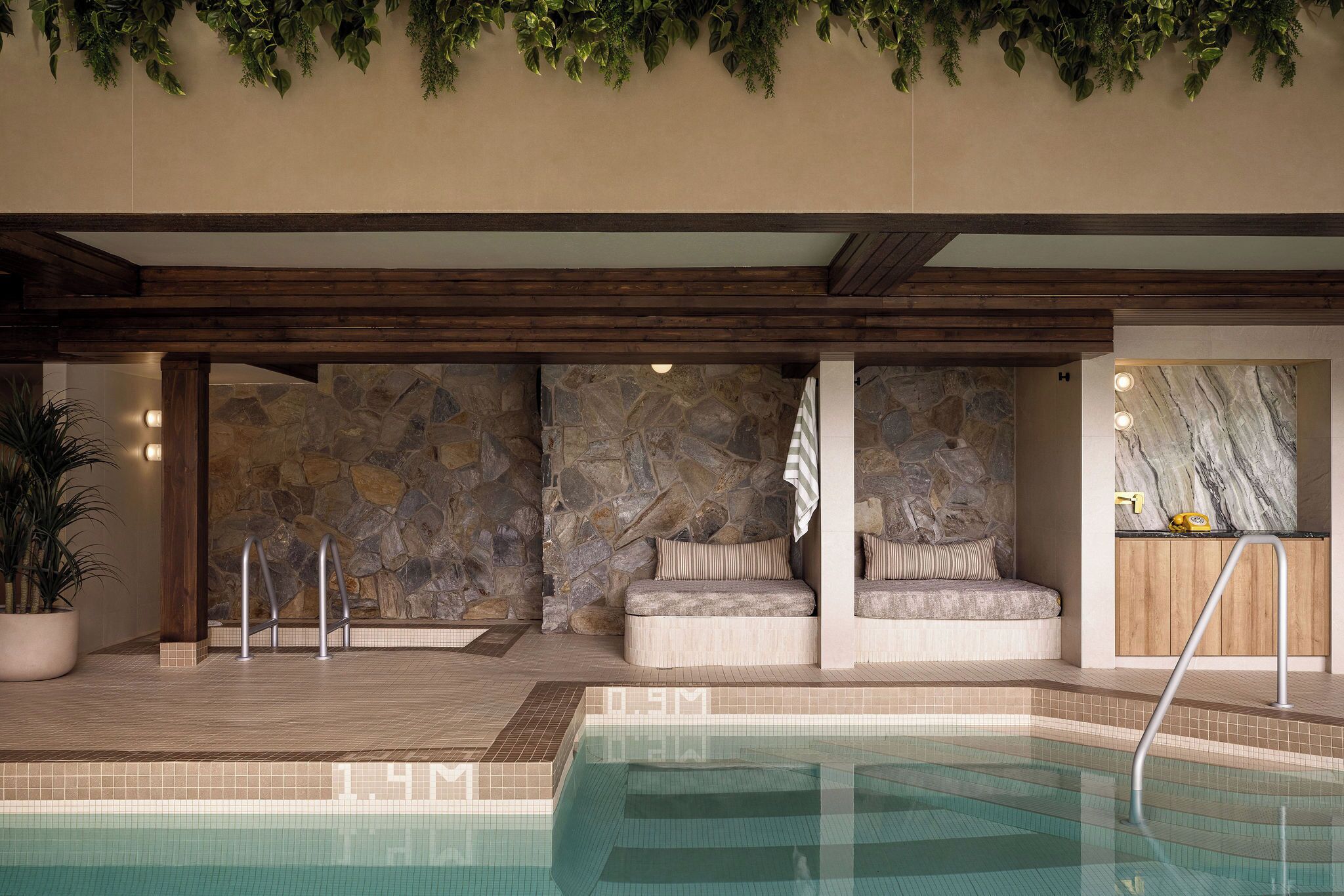 Zona de piscina con muros de piedra, techo de madera y área de descanso con cojines.