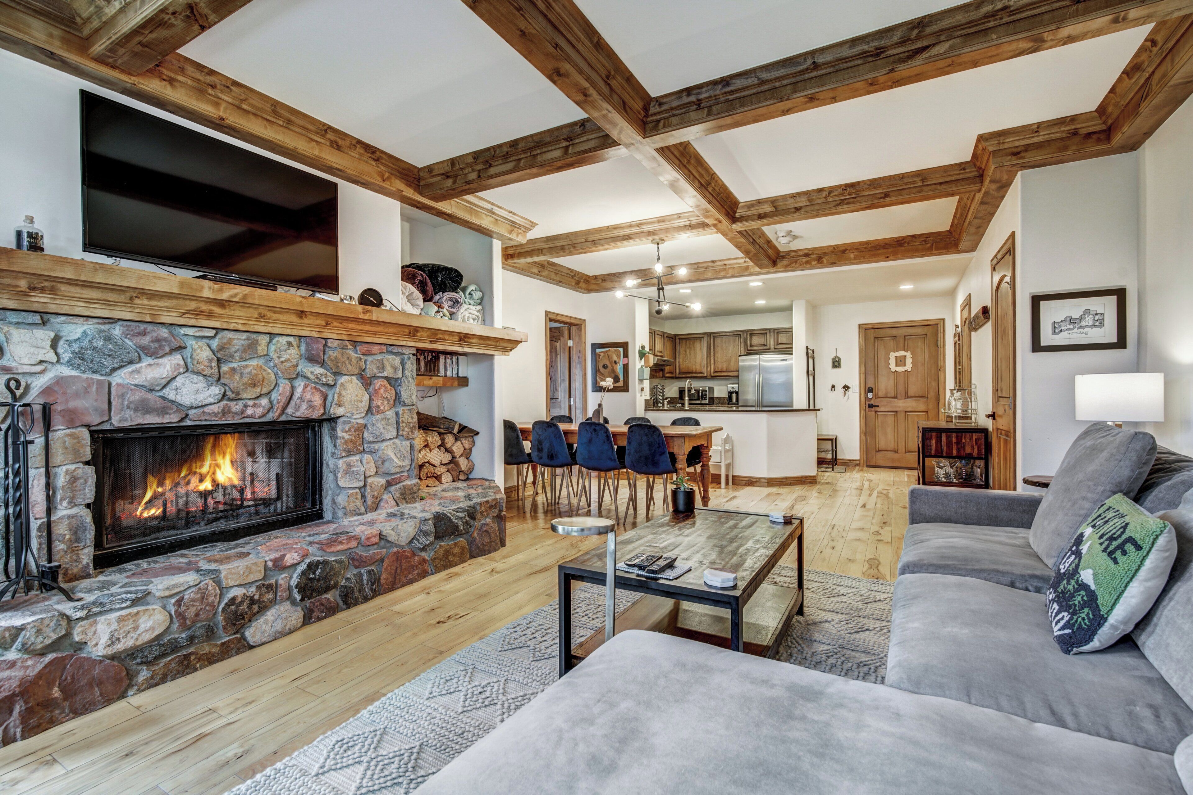 Ein gemütliches Wohnzimmer mit Kamin aus Stein, Holzbalken, einem grauen Sofa und einem Essbereich mit Stühlen.