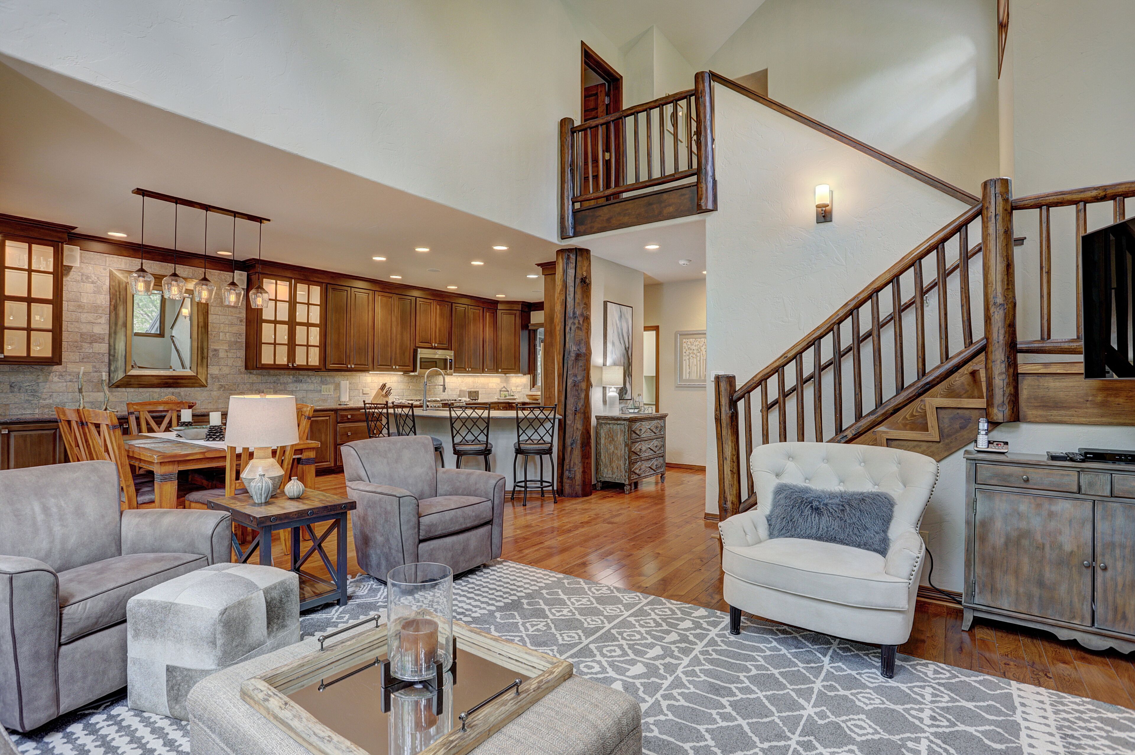 A spacious living area with a high ceiling, wooden staircase, and a kitchen in the background.