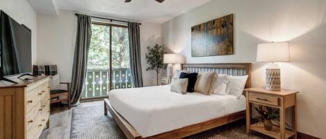 Un dormitorio con cama de madera, ventilador de techo, ventana grande con cortinas, mesita de noche con lámpara y un cuadro en la pared.