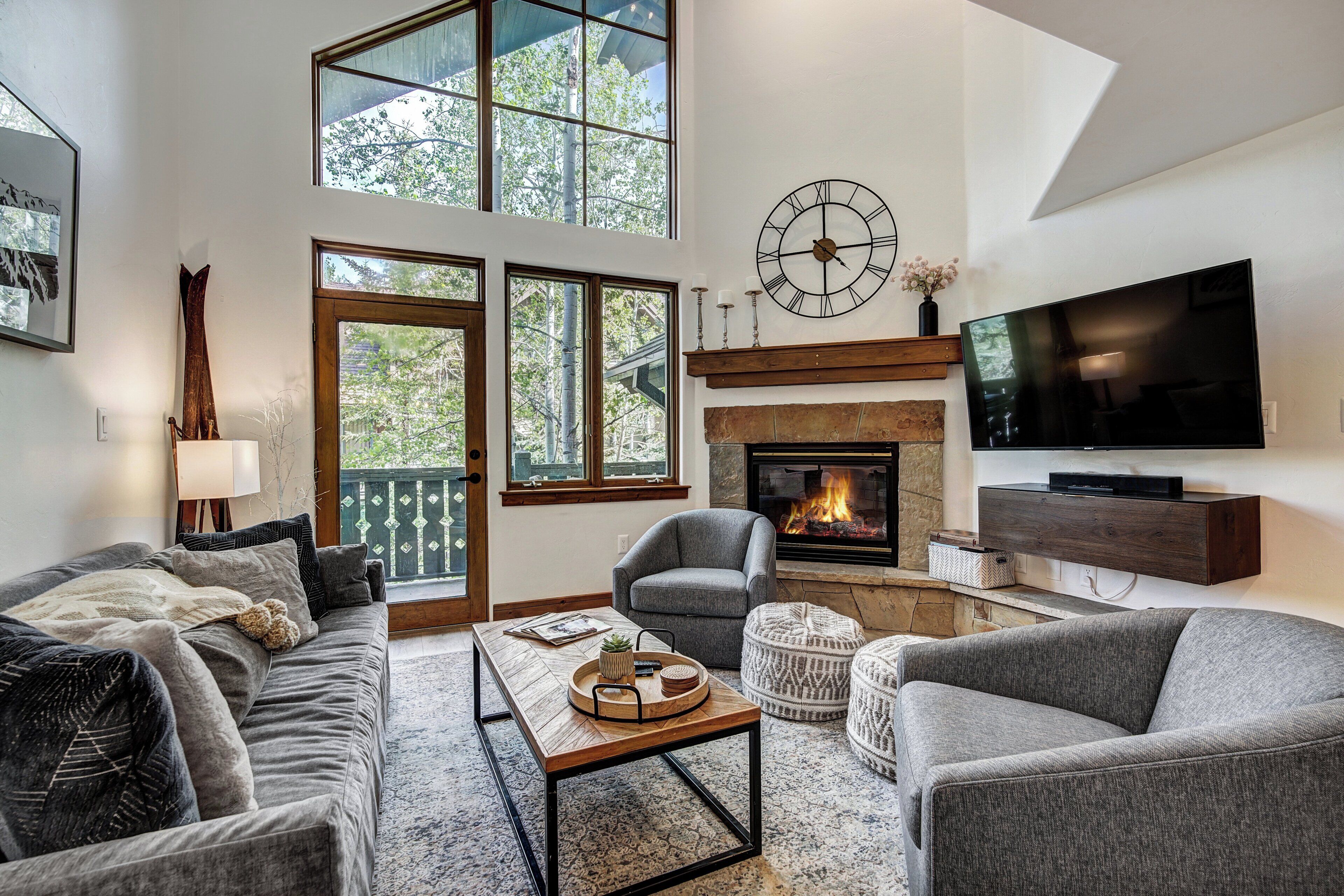 A modern living room with a fireplace, a television, and a large window overlooking a deck.