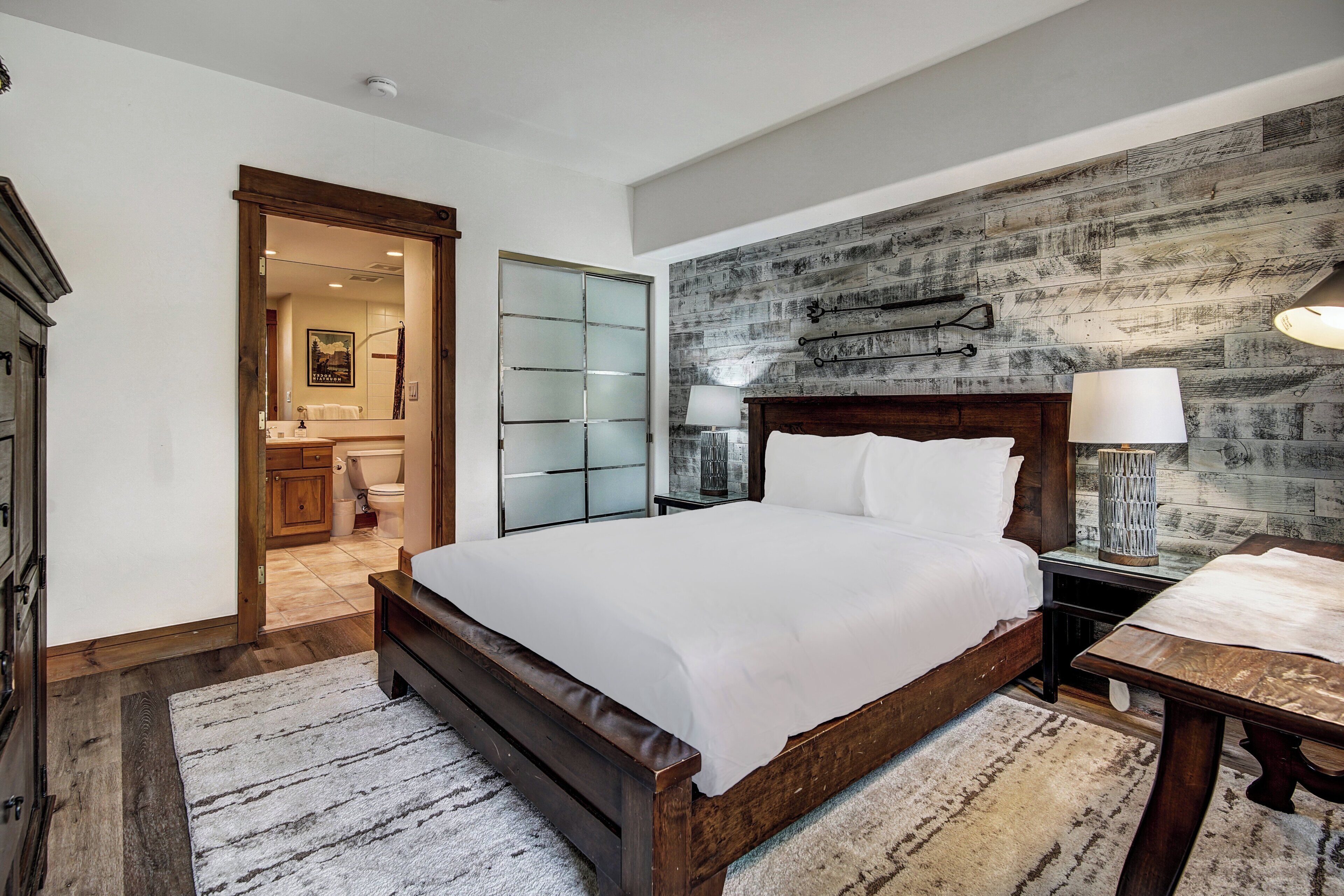 A bedroom with a wooden bed, a nightstand with a lamp, a wooden headboard, and a large window with a view of the bathroom.