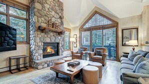 A spacious living room with a stone fireplace, comfortable seating, and large windows offering a view of trees. - Arrowhead Village, A Vail Resorts Property (Edwards)