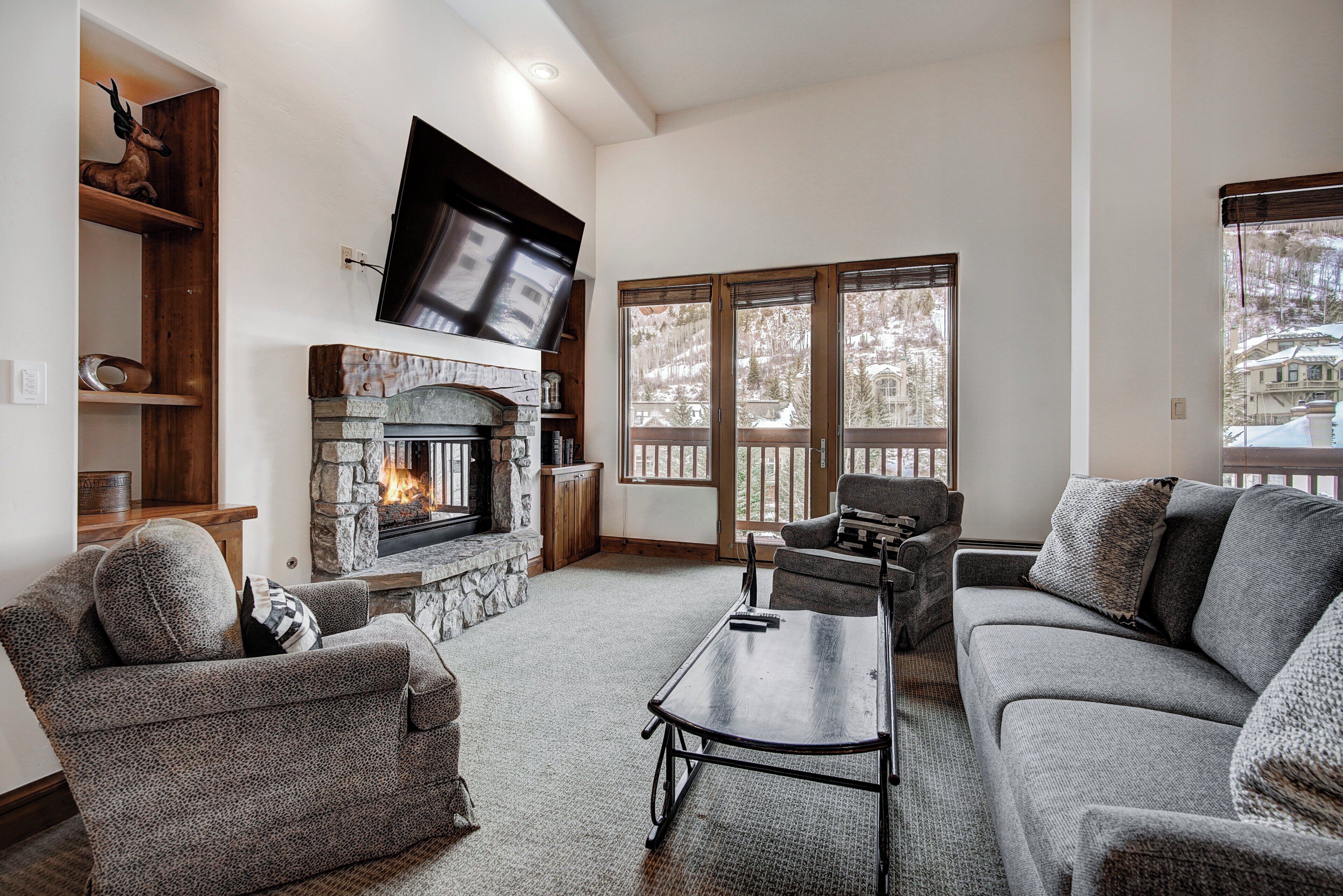 A cozy living room with a stone fireplace, a flat-screen TV mounted above it, and a large window offering a view of a snowy landscape.
