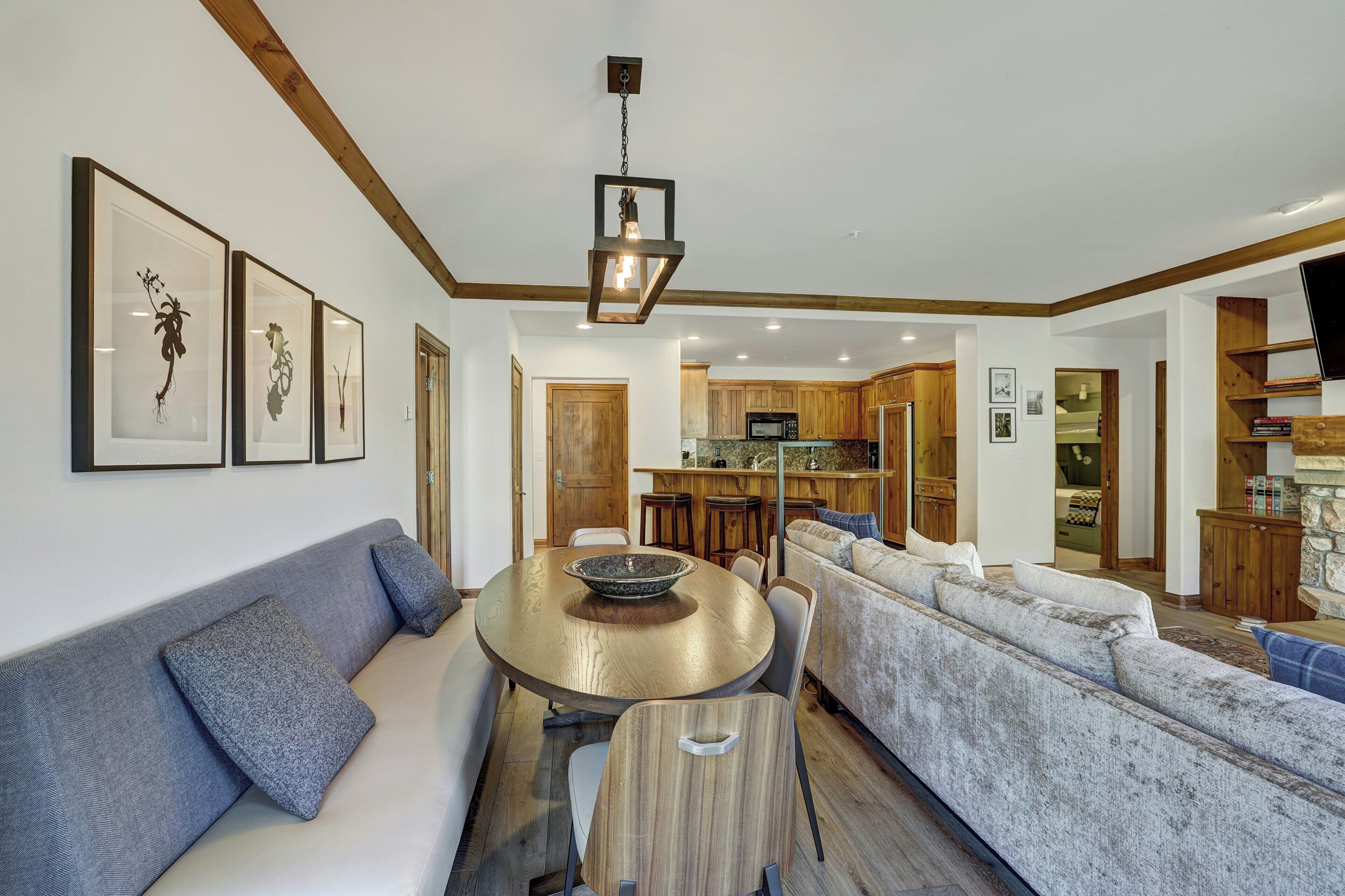 A modern living room with a round wooden table, a grey sofa, and a large grey sectional sofa. There is a kitchen in the background with wooden cabinets and a microwave.