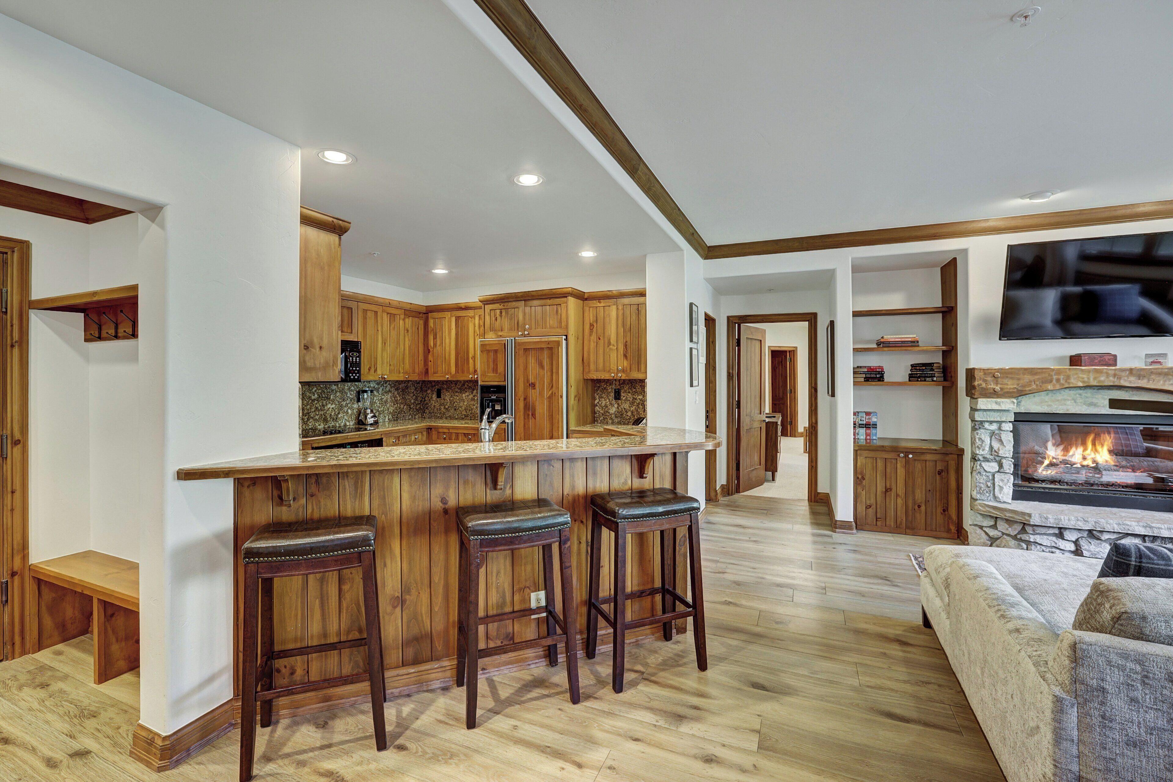 A modern kitchen with wooden cabinets, a central island with bar stools, and a stone fireplace.
