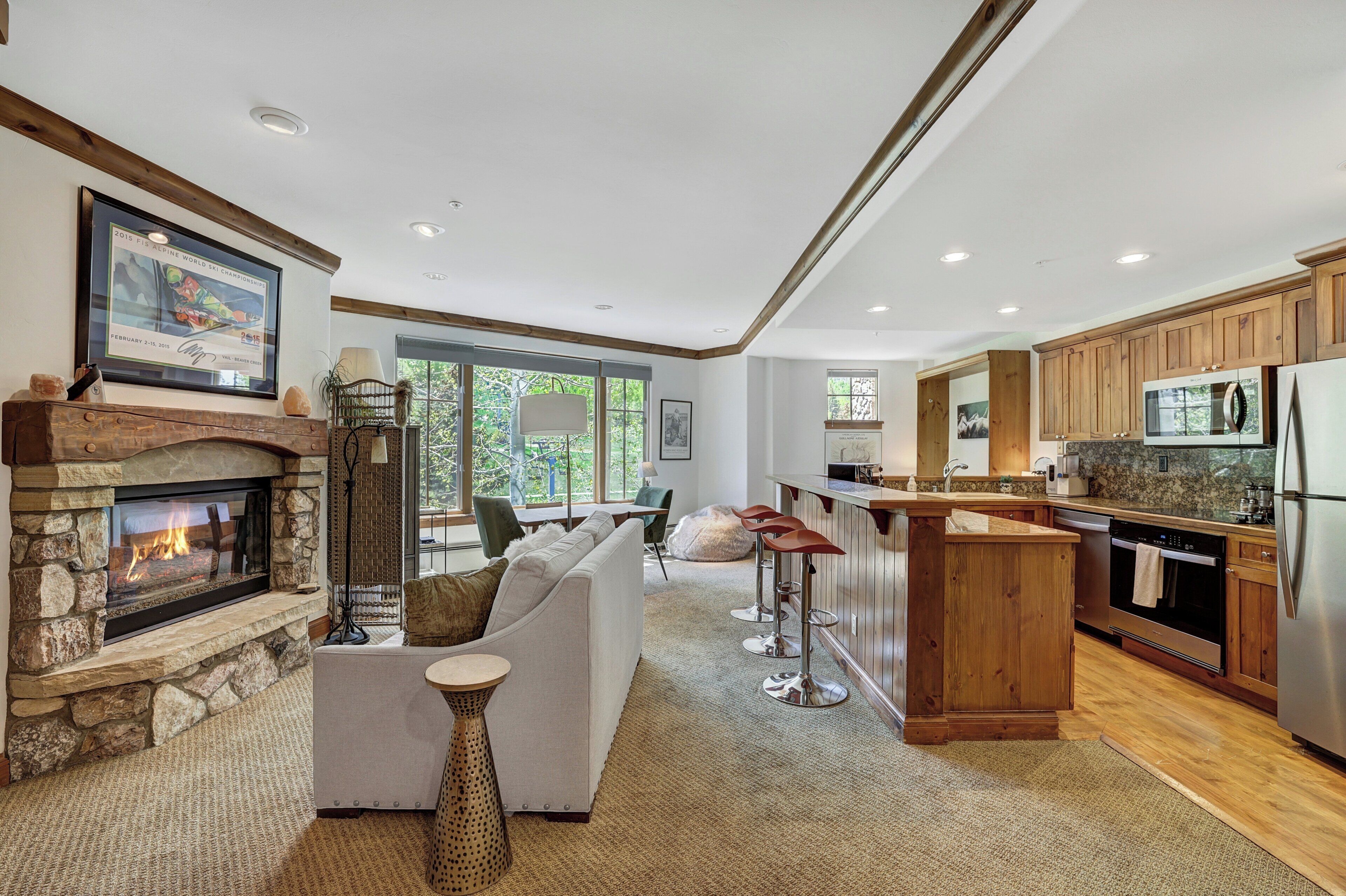 A spacious living room with a stone fireplace, a white sofa, and a kitchen area with wooden cabinets and stainless steel appliances.