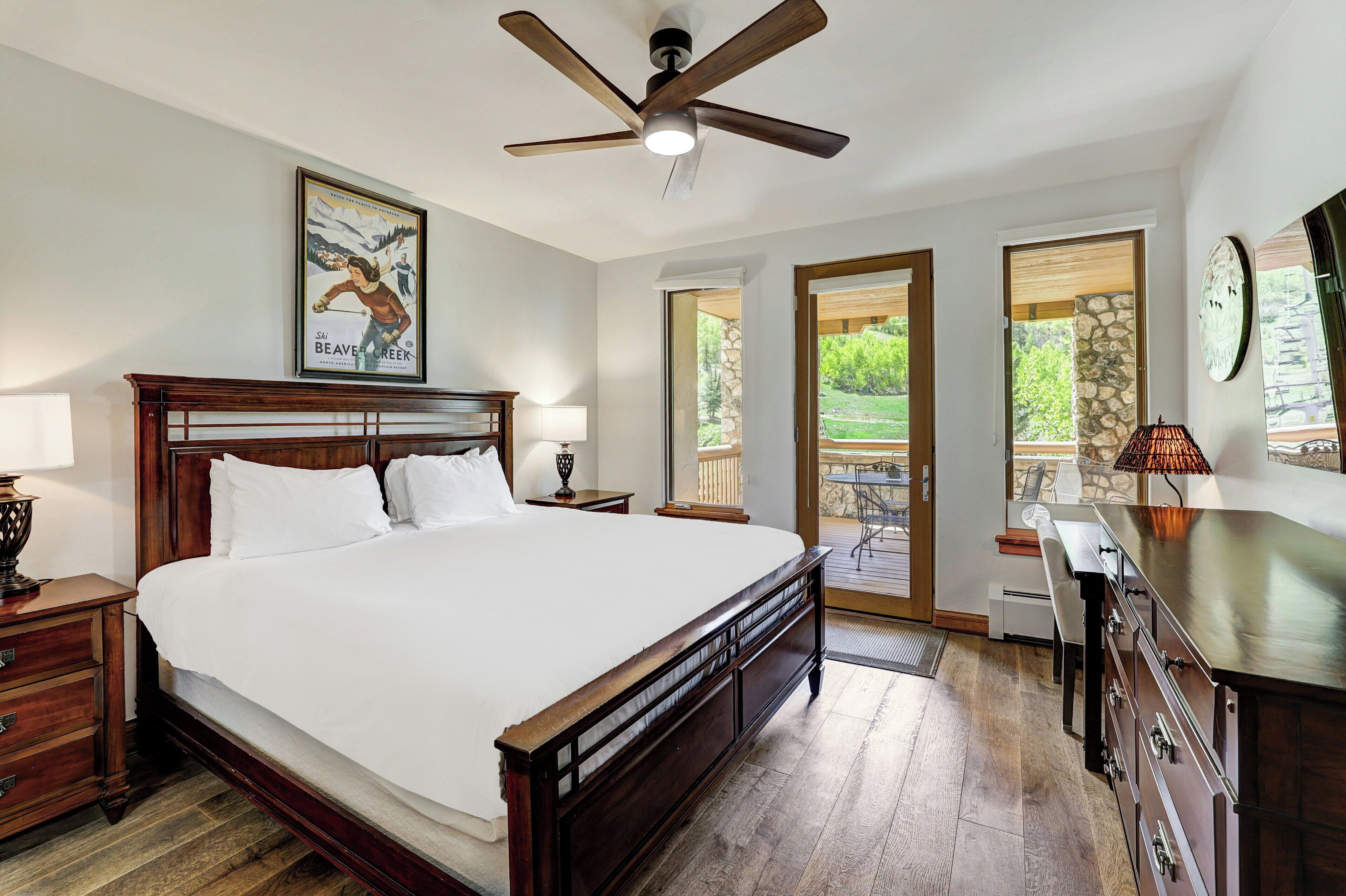A bedroom with a large bed, a ceiling fan, a wooden nightstand, a dresser, and a view of the outdoors through a glass door.