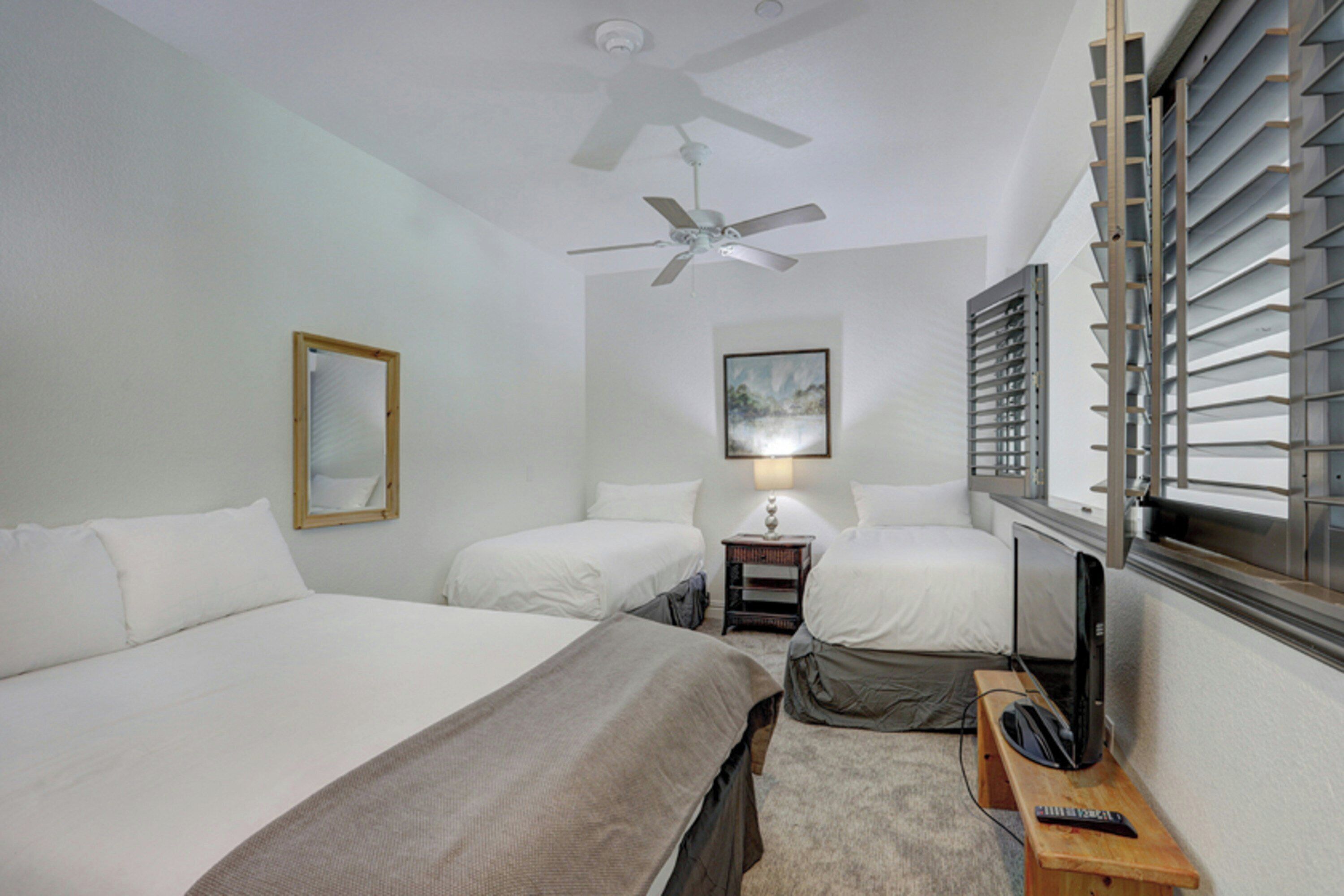 A hotel room with two single beds, a ceiling fan, a bedside table with a lamp, a TV, and a window with blinds.