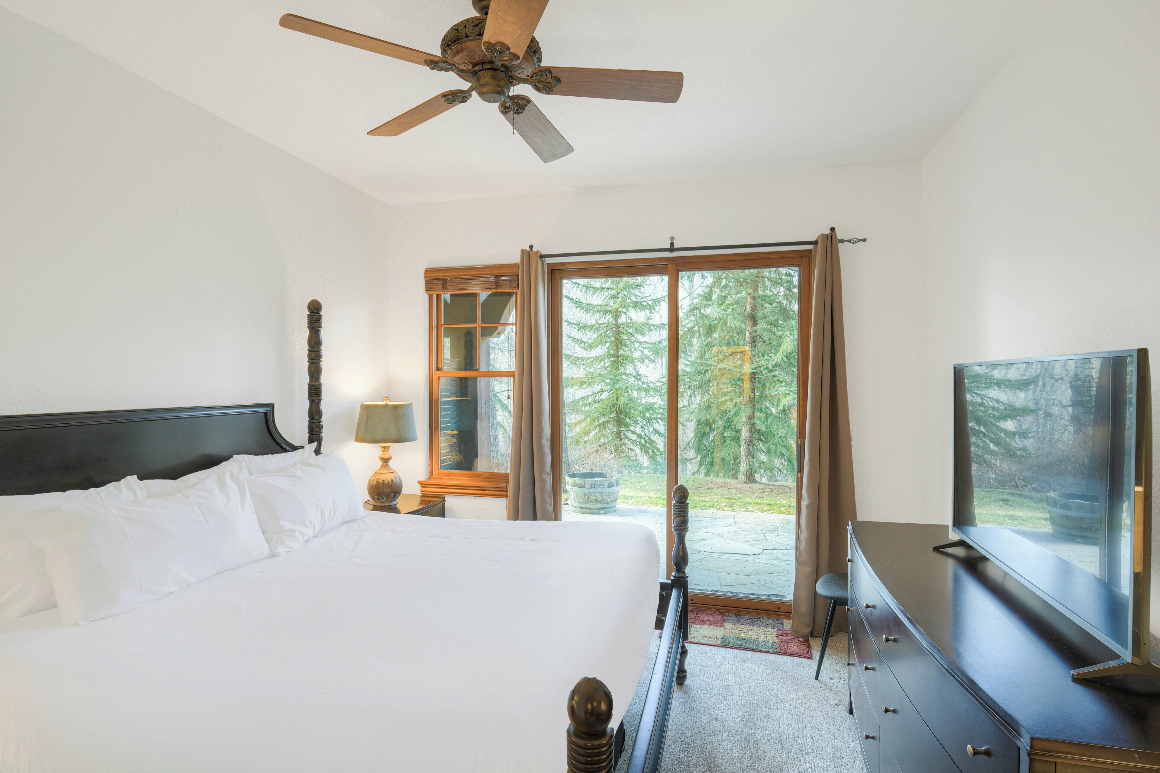 A bedroom with a bed, a ceiling fan, a dresser, a television, and a large window with a view of trees.