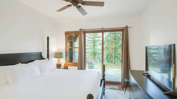 Un dormitorio con cama, ventilador de techo, cómoda, televisor y un amplio ventanal con vista a árboles.