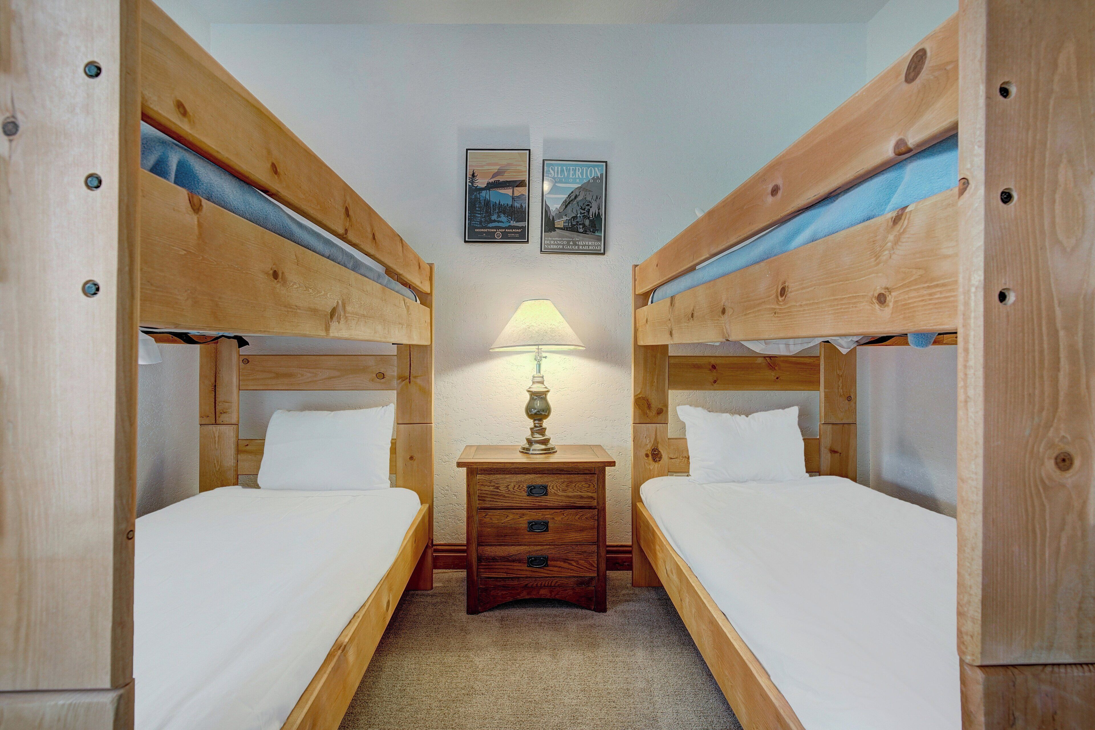 A room with two wooden bunk beds, a small wooden nightstand with a lamp, and two framed pictures on the wall.