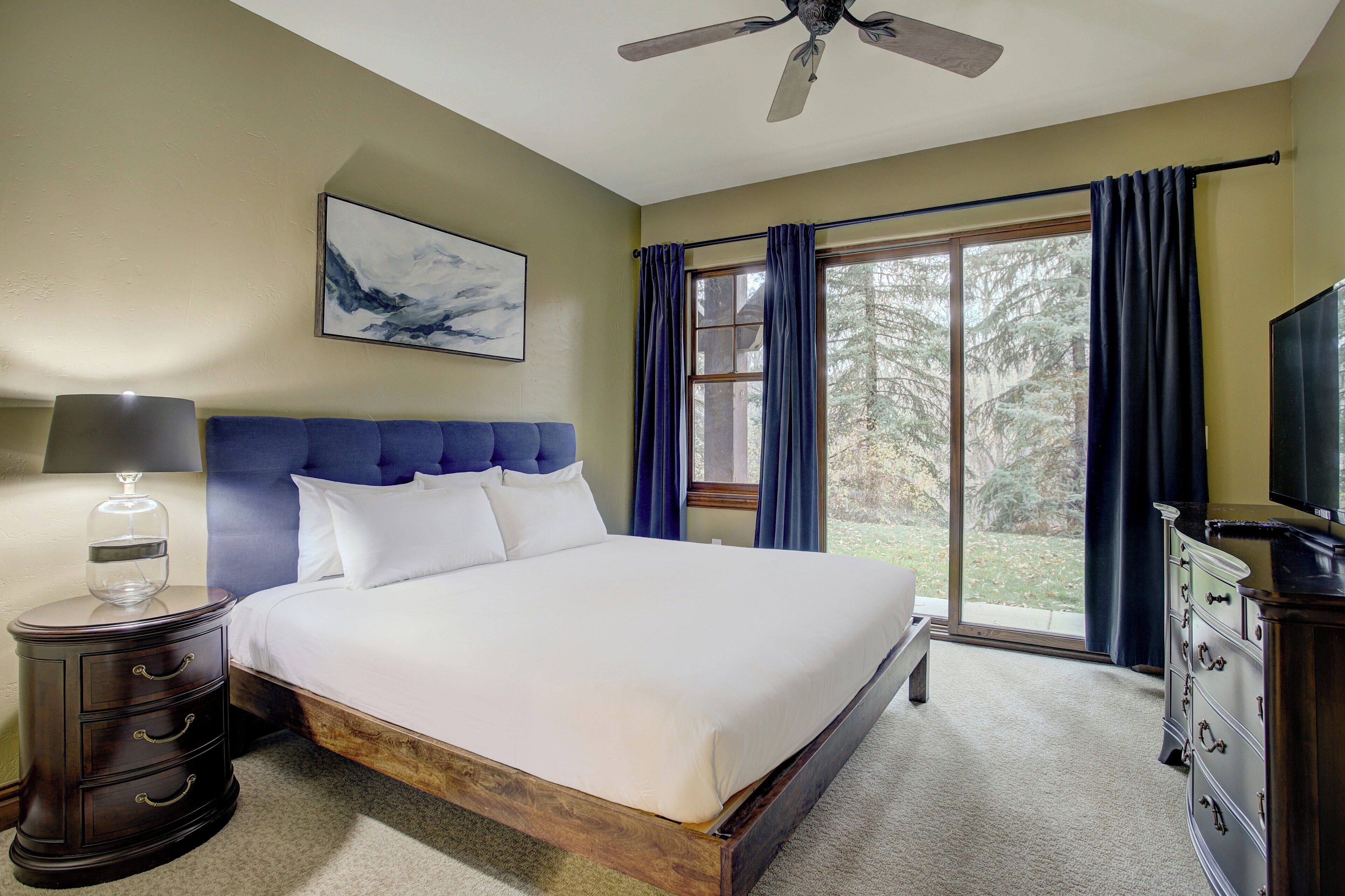 A bedroom with a bed, a nightstand, a dresser, a ceiling fan, a window with curtains, and a painting on the wall.
