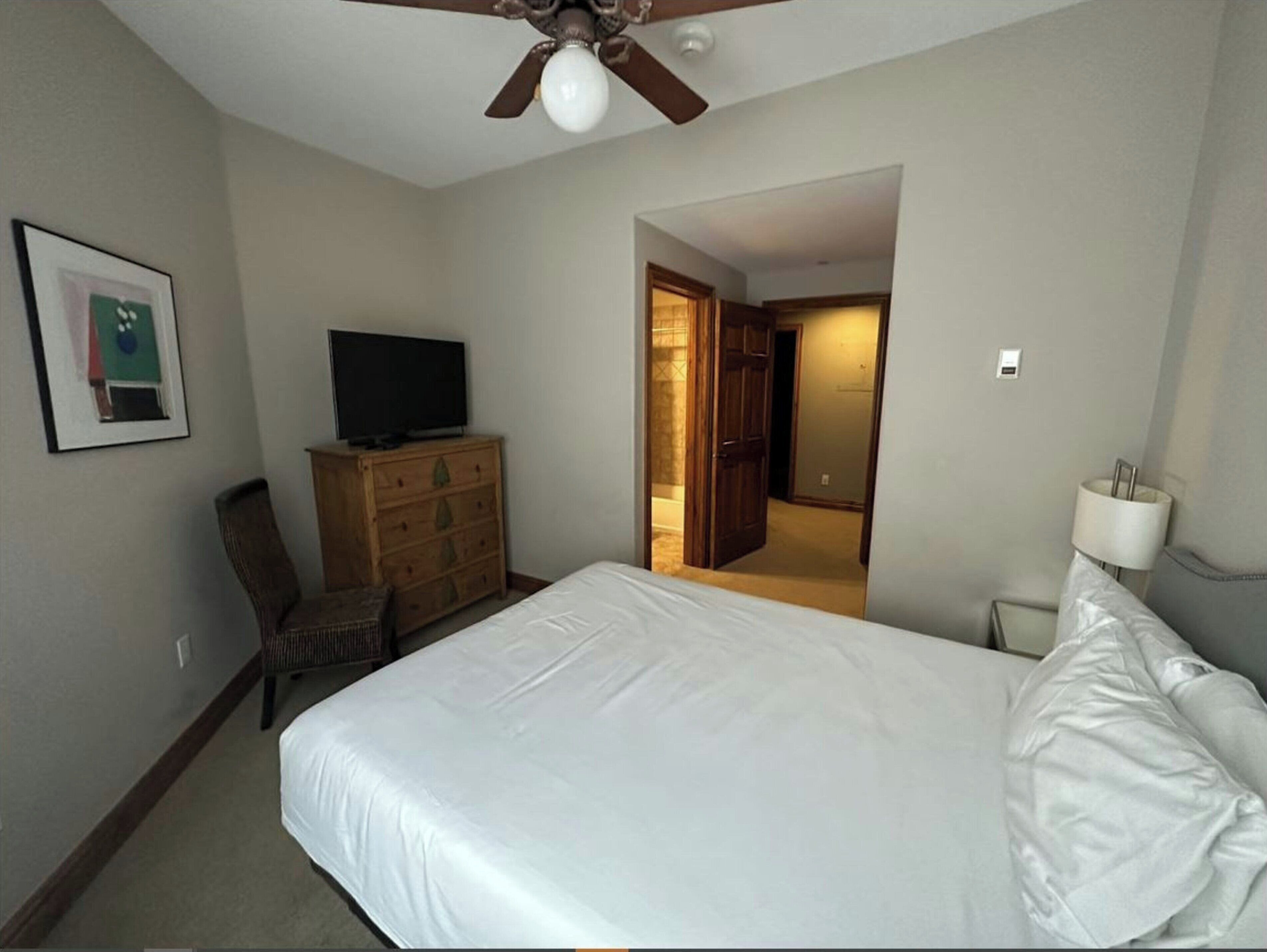 A hotel room with a bed, a television on a dresser, a chair, and a door leading to another room.