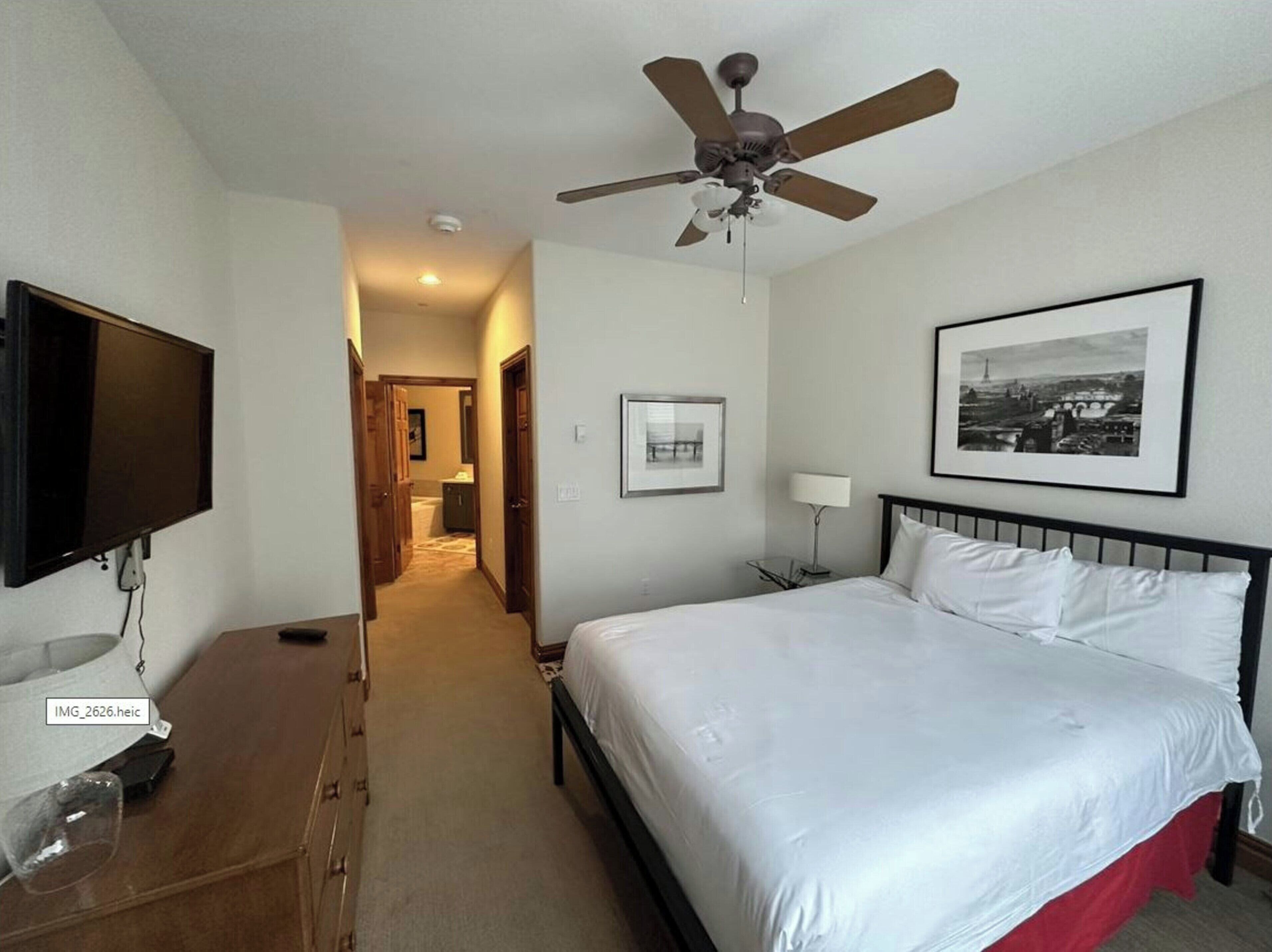 Una habitación de hotel con una cama, un ventilador de techo, un televisor y un cuadro en la pared.