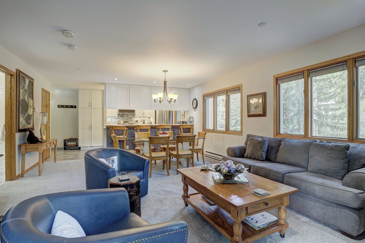 A spacious living area with a grey sofa, blue armchair, and wooden coffee table. A kitchen with white cabinets and a dining area with wooden chairs is visible in the background.