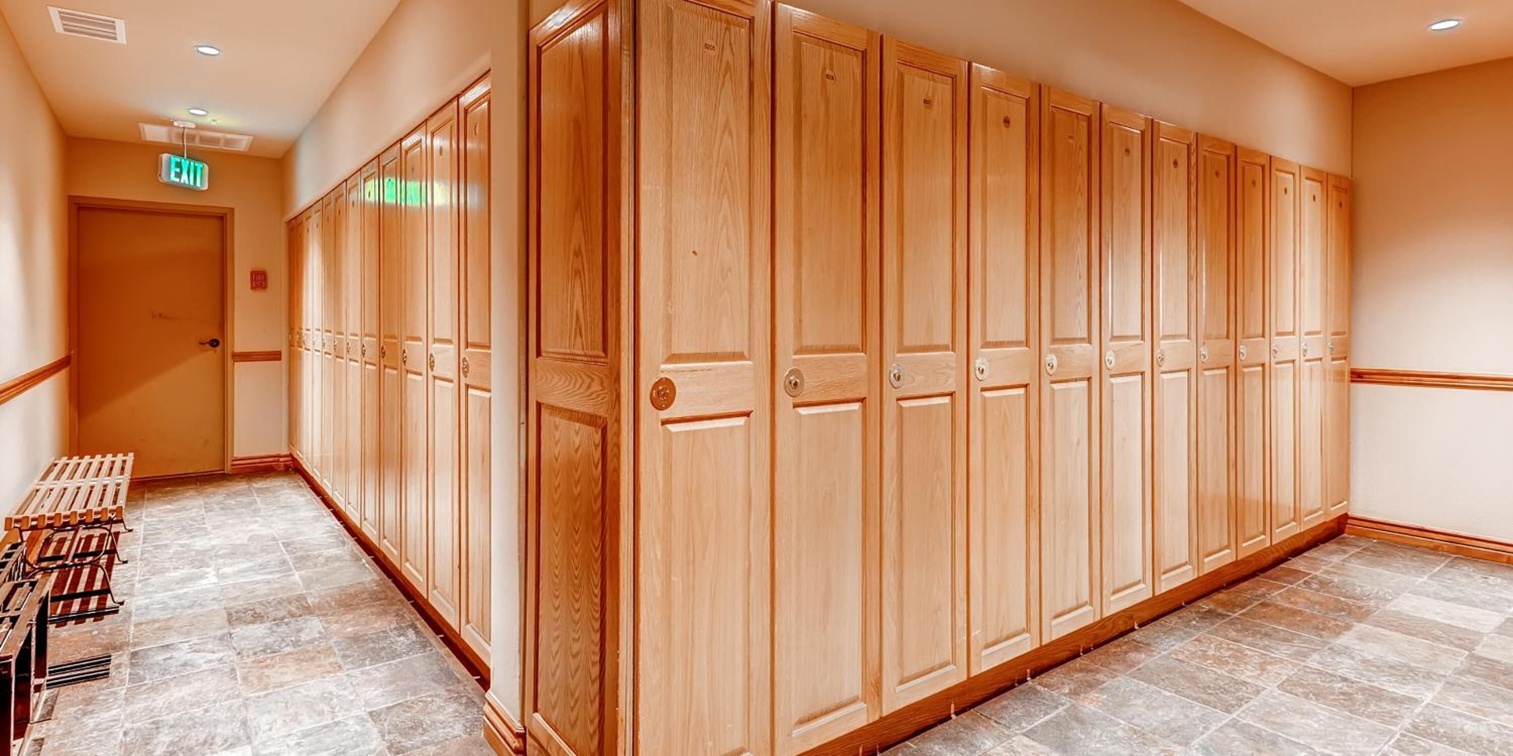 A hallway with wooden lockers, a bench, and an exit sign.