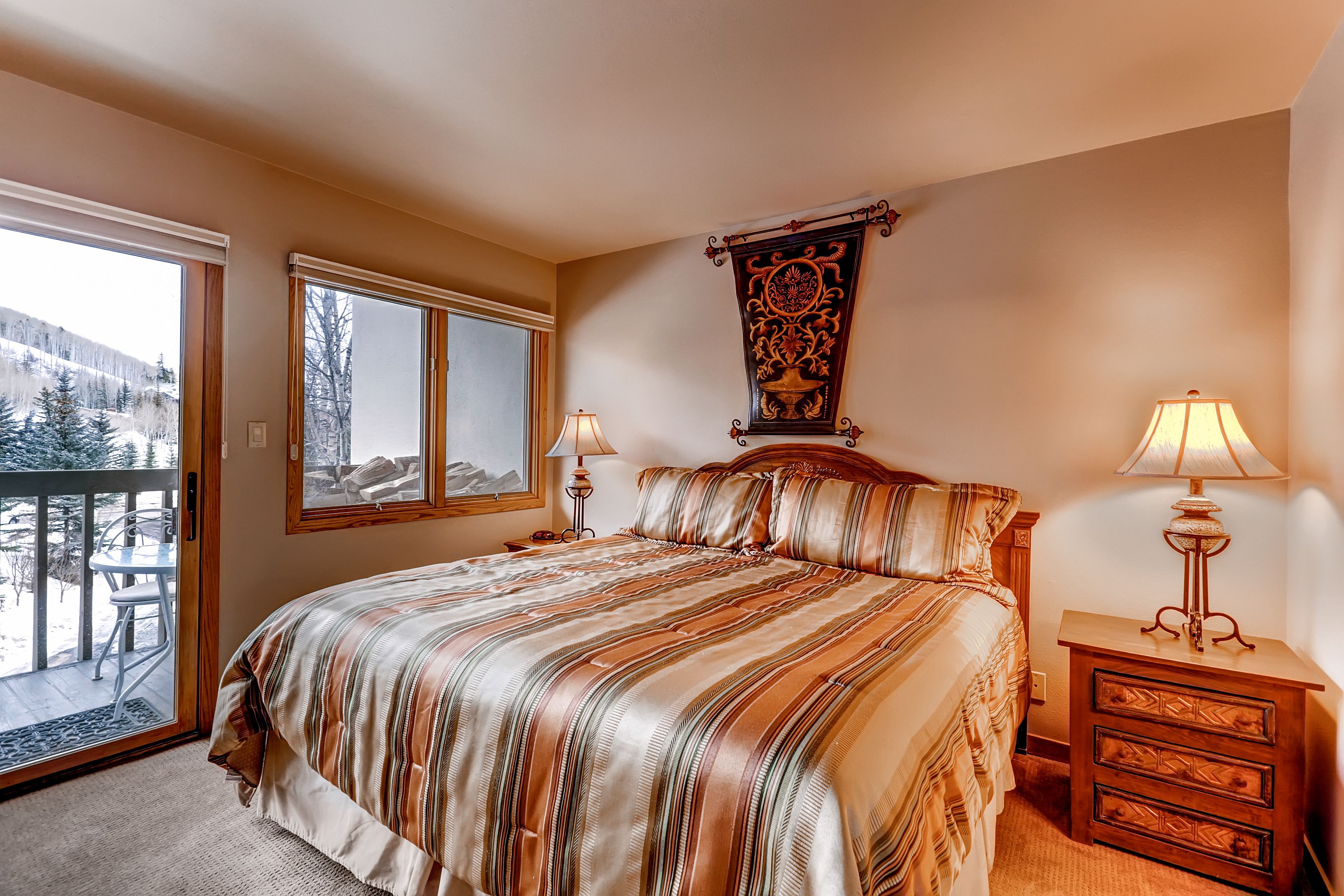 A bedroom with a large bed, two bedside tables, a wooden nightstand, a decorative wall hanging, and a view of a snowy landscape through a sliding door.
