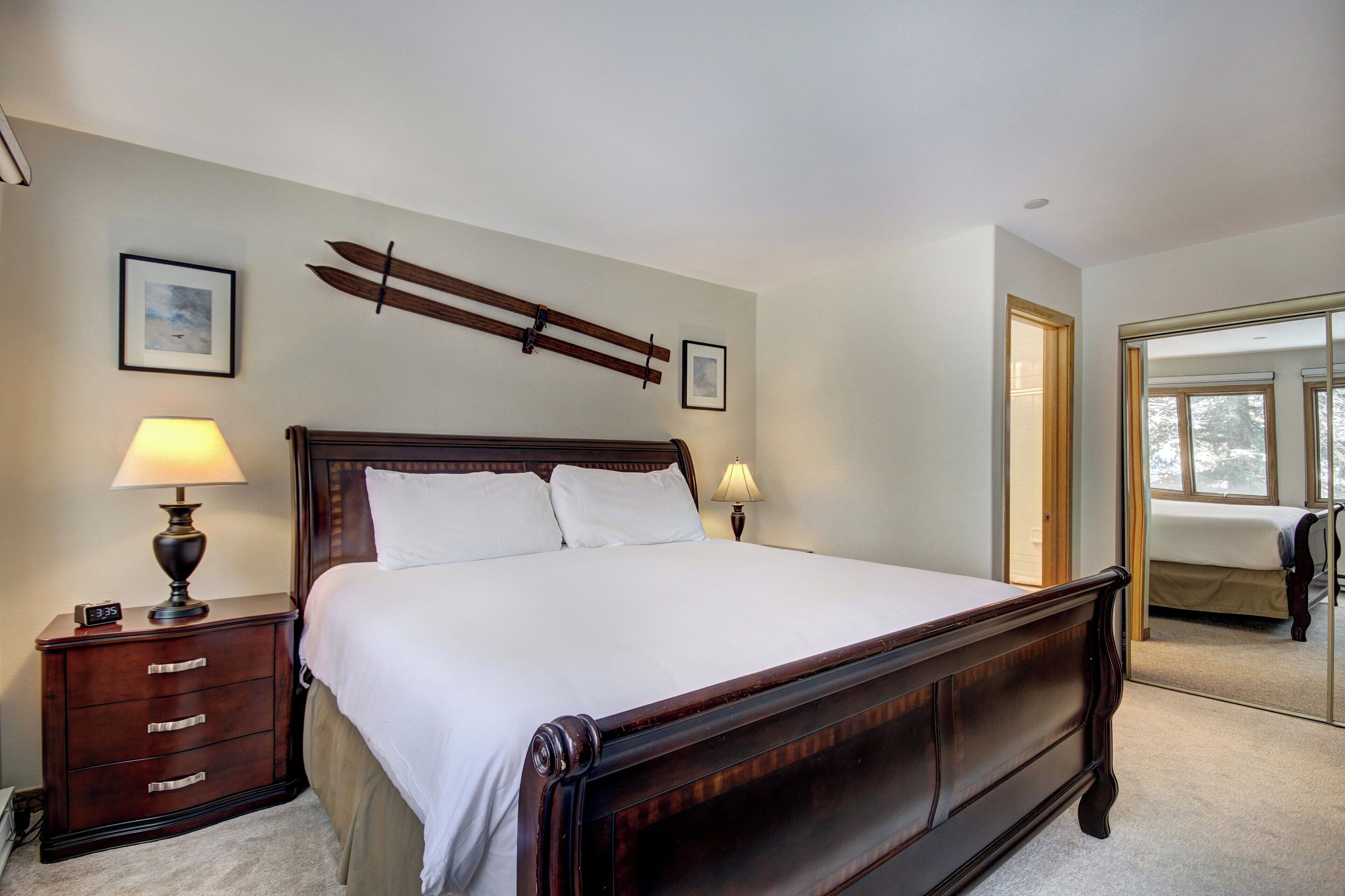 A neatly made bed with a wooden headboard, two bedside lamps, a nightstand with drawers, and a framed picture on the wall.