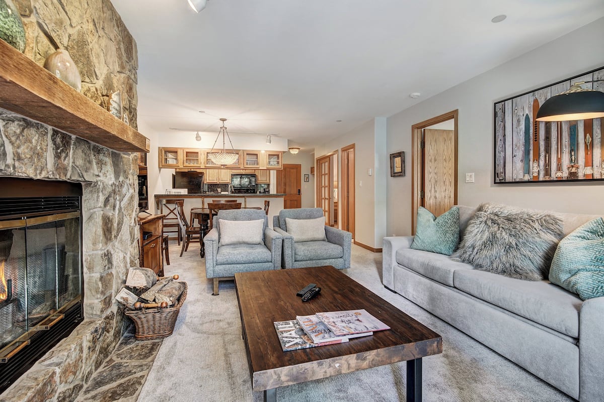 A cozy living room with a fireplace, a coffee table, and a sofa. There is a kitchen in the background with wooden cabinets and a dining area.