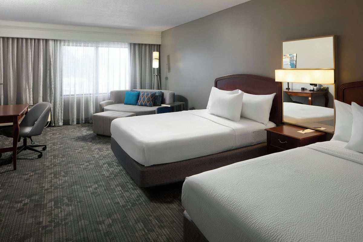 A hotel room with two beds, a desk, a chair, and a lamp.