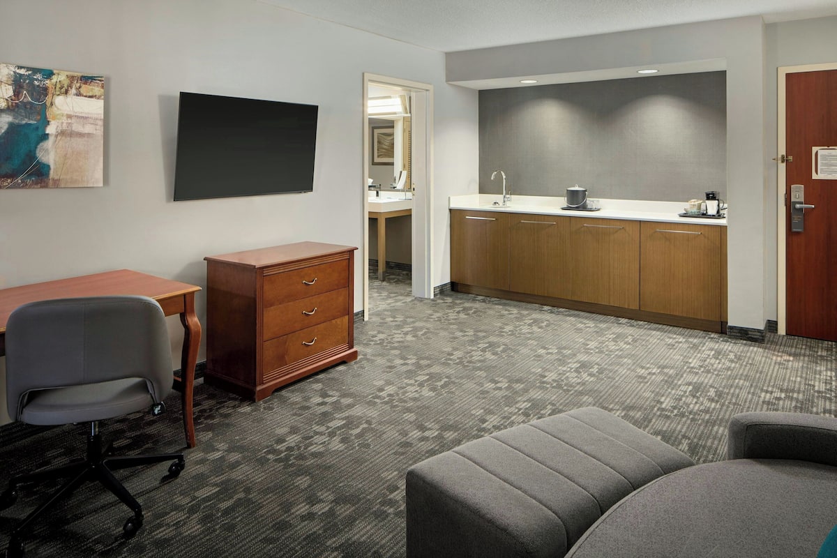 A hotel room with a desk, a chair, a dresser, a kitchenette, and a large window.