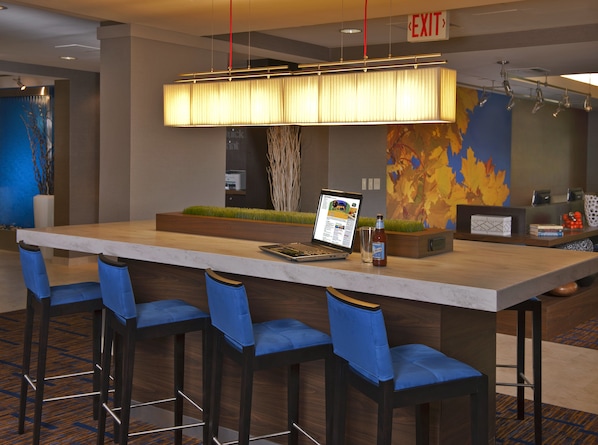 A modern hotel lobby with a long reception desk, blue upholstered bar stools, a laptop, and a bottle of Moon beer.