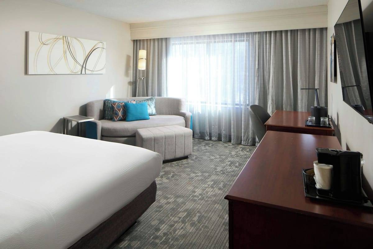 A hotel room with a large bed, a desk with a coffee maker, a sofa with pillows, a TV, and a large window with curtains.