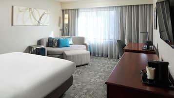 Habitación de hotel con una cama grande, un escritorio con cafetera, un sofá con almohadas, un televisor y un ventanal con cortinas.