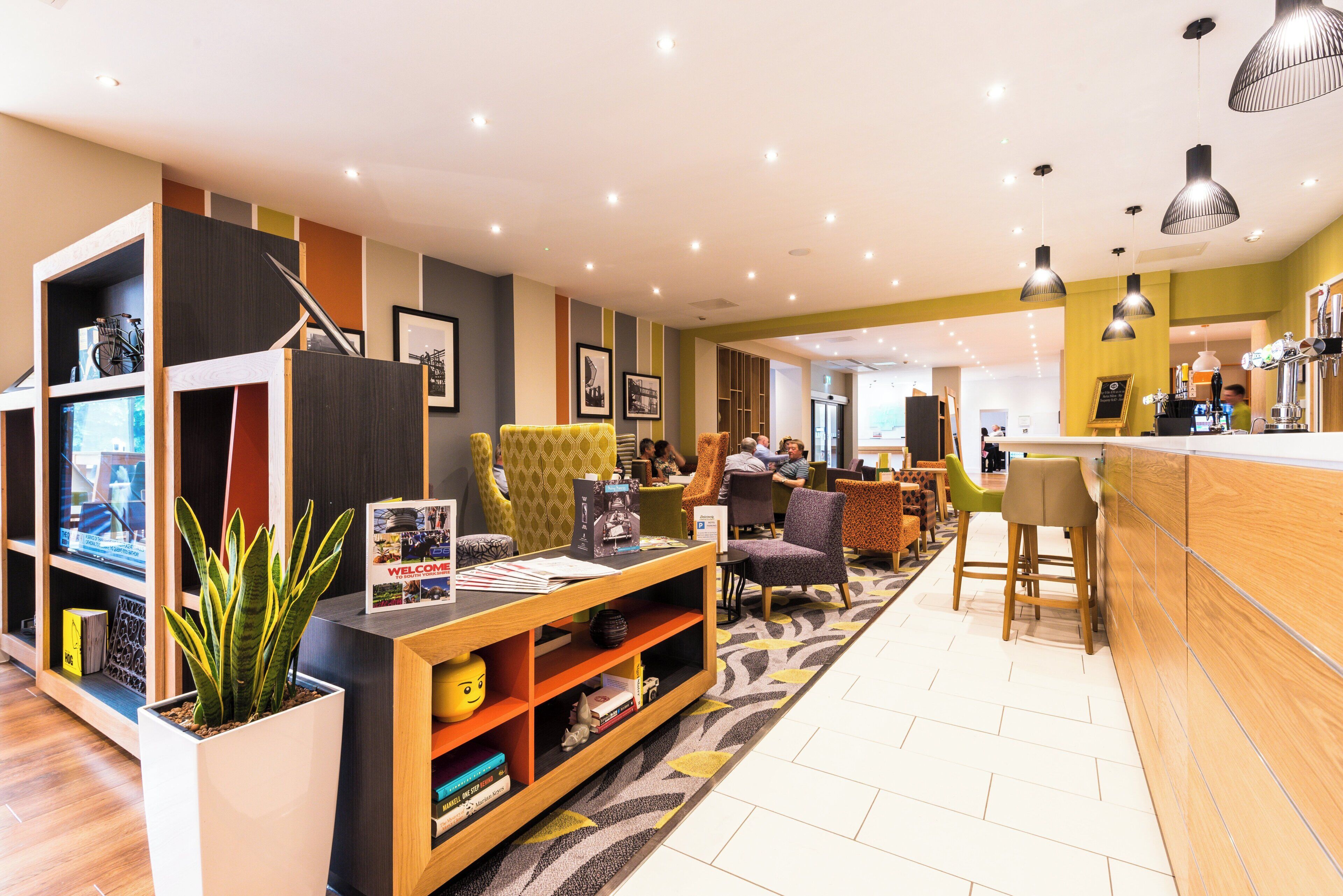 A modern lounge area with seating, a coffee bar, and a bookshelf with books and a plant.