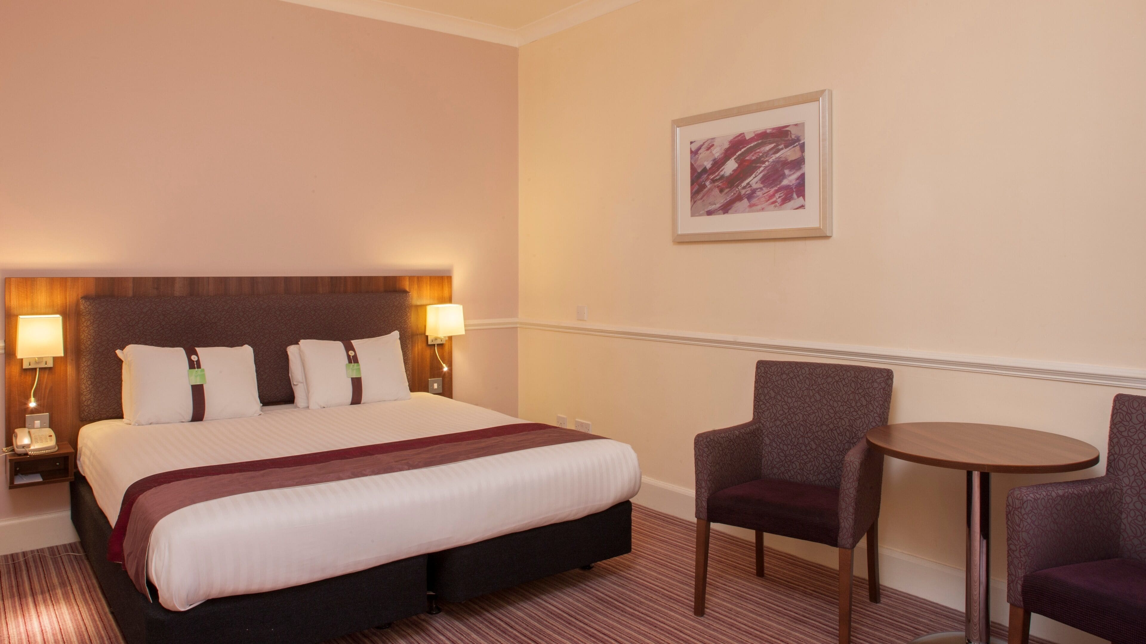 Una habitación de hotel con una cama grande, dos lámparas de noche, una mesita, dos sillas y un cuadro en la pared.