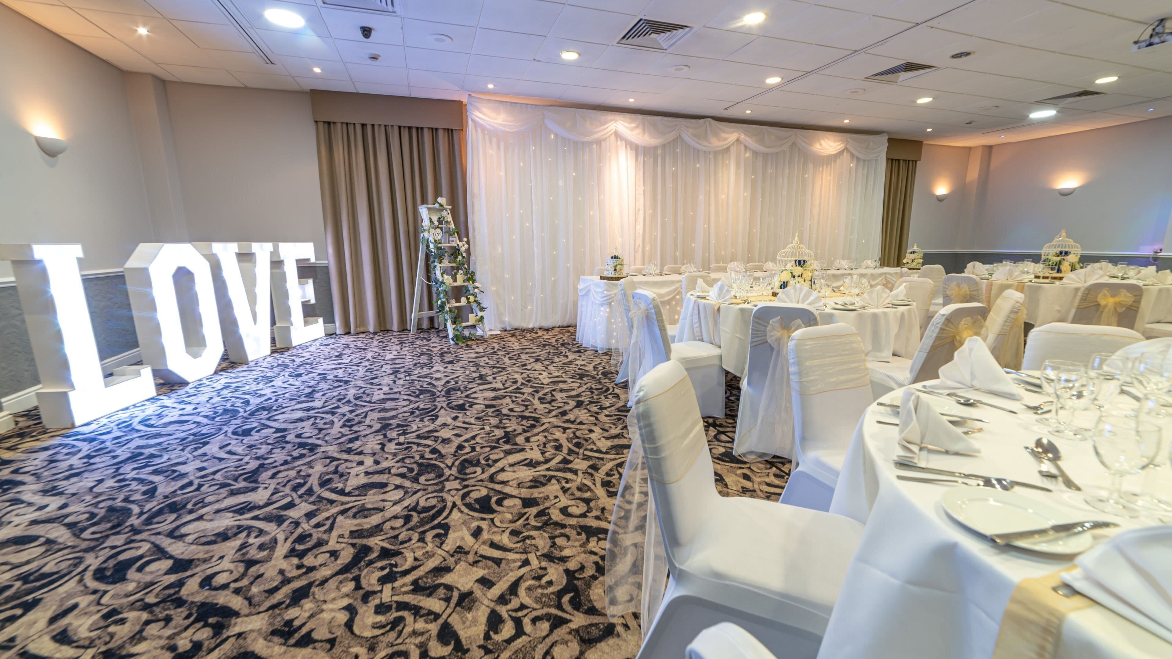 Een balzaal met ronde tafels gedekt voor een formeel evenement, een groot 'LOVE' bord en een bloemstuk.