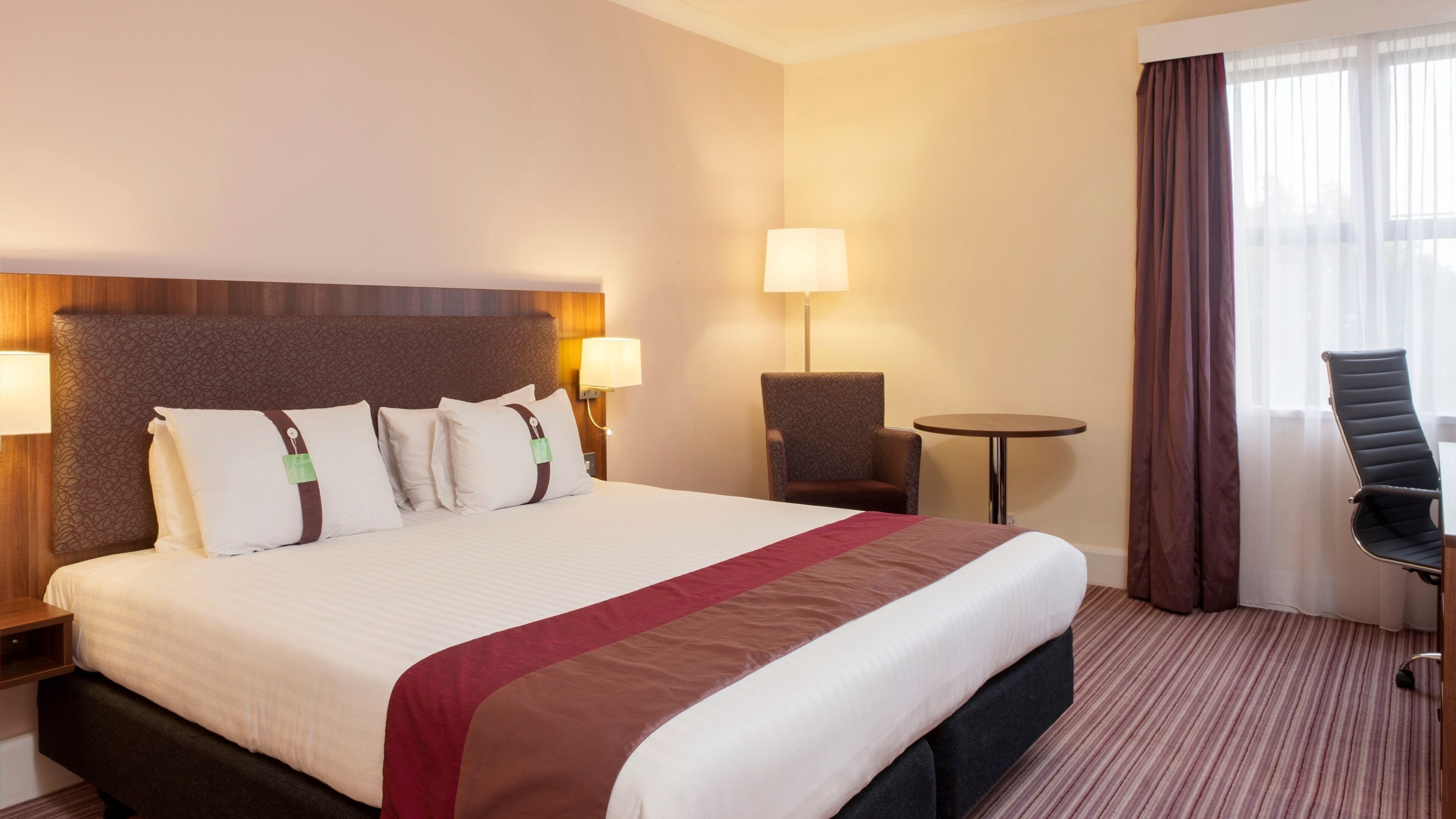A hotel room with a large bed, two bedside lamps, a small table, a chair, and a view of the outdoors through sheer curtains.