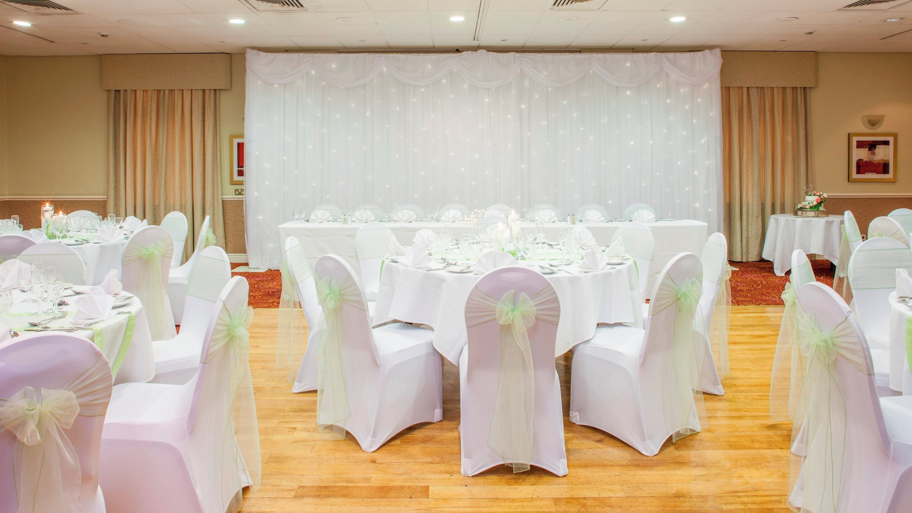 Une salle de banquet avec des tables rondes dressées pour le repas, des chaises recouvertes de housses blanches et une scène ornée de rideaux.
