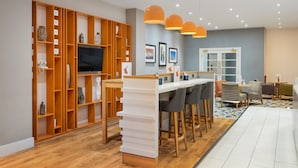 A modern living room with a wooden bookshelf, a flat-screen TV, a white bar area, and a seating area with a patterned rug. - Holiday Inn Rotherham Sheffield by IHG (Rotherham)
