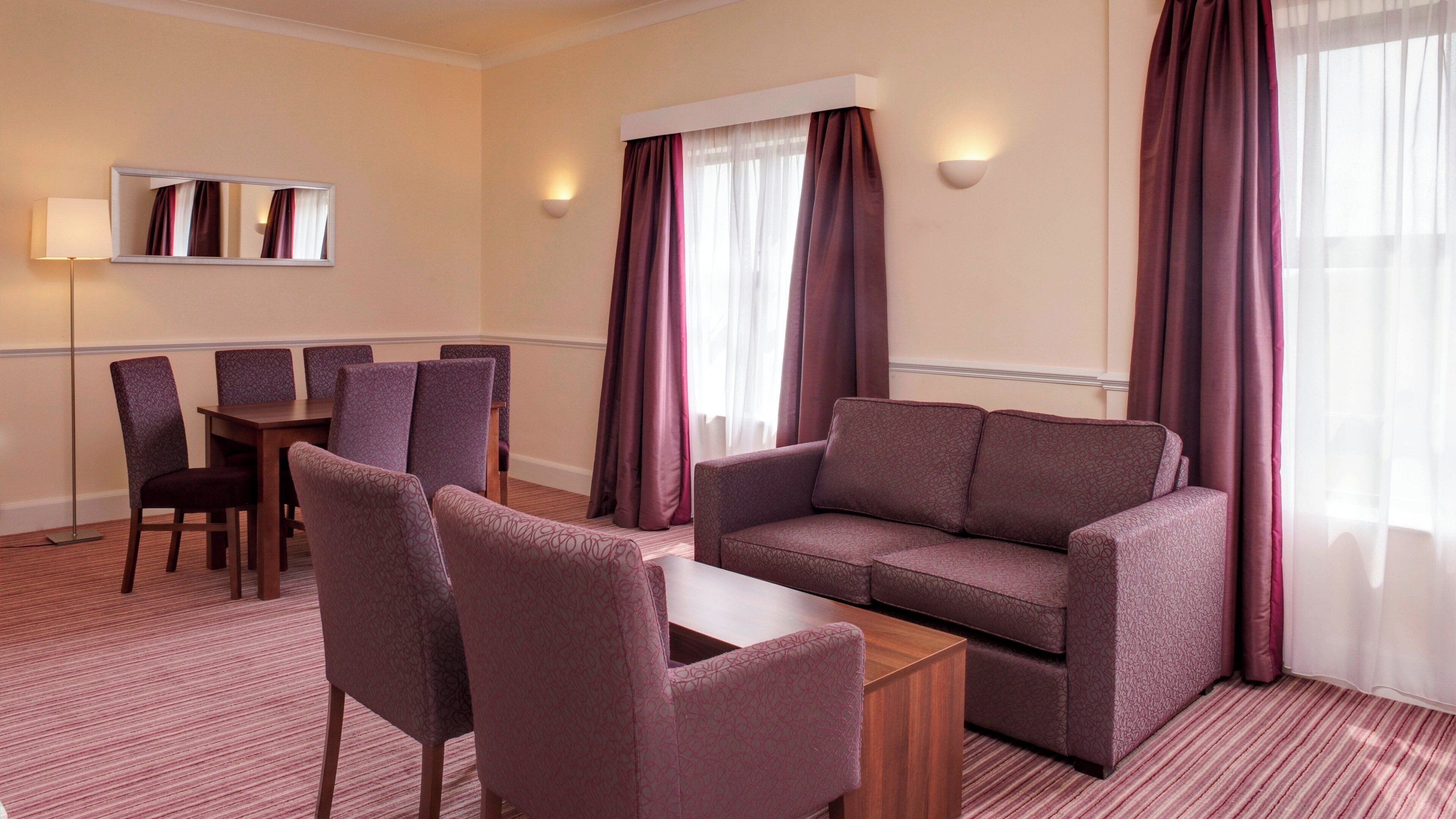 A hotel room with a sofa, chairs, a small table, a lamp, and a mirror.