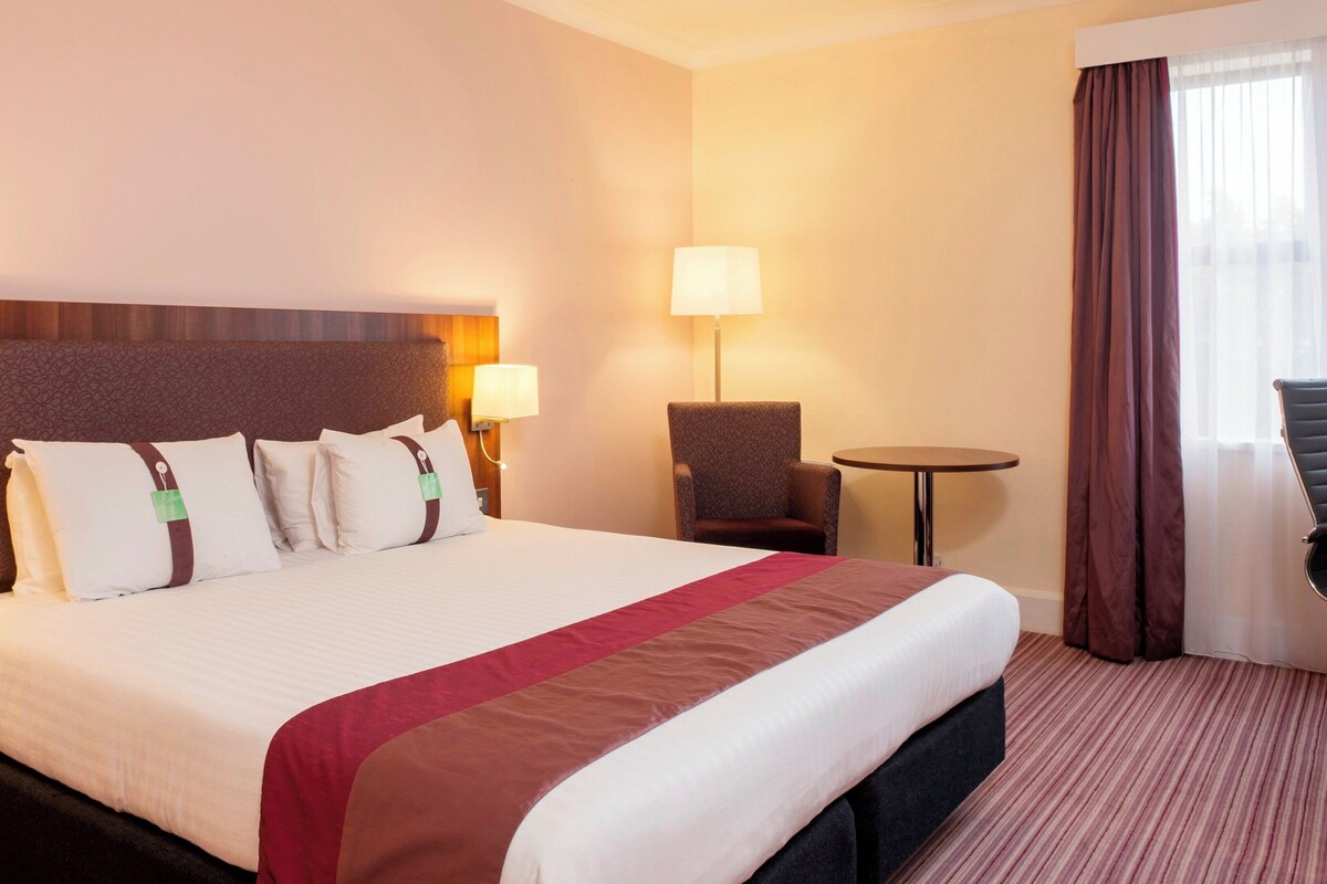 A hotel room with a large bed, two bedside lamps, a small round table, a chair, and a view of the outdoors through the window.