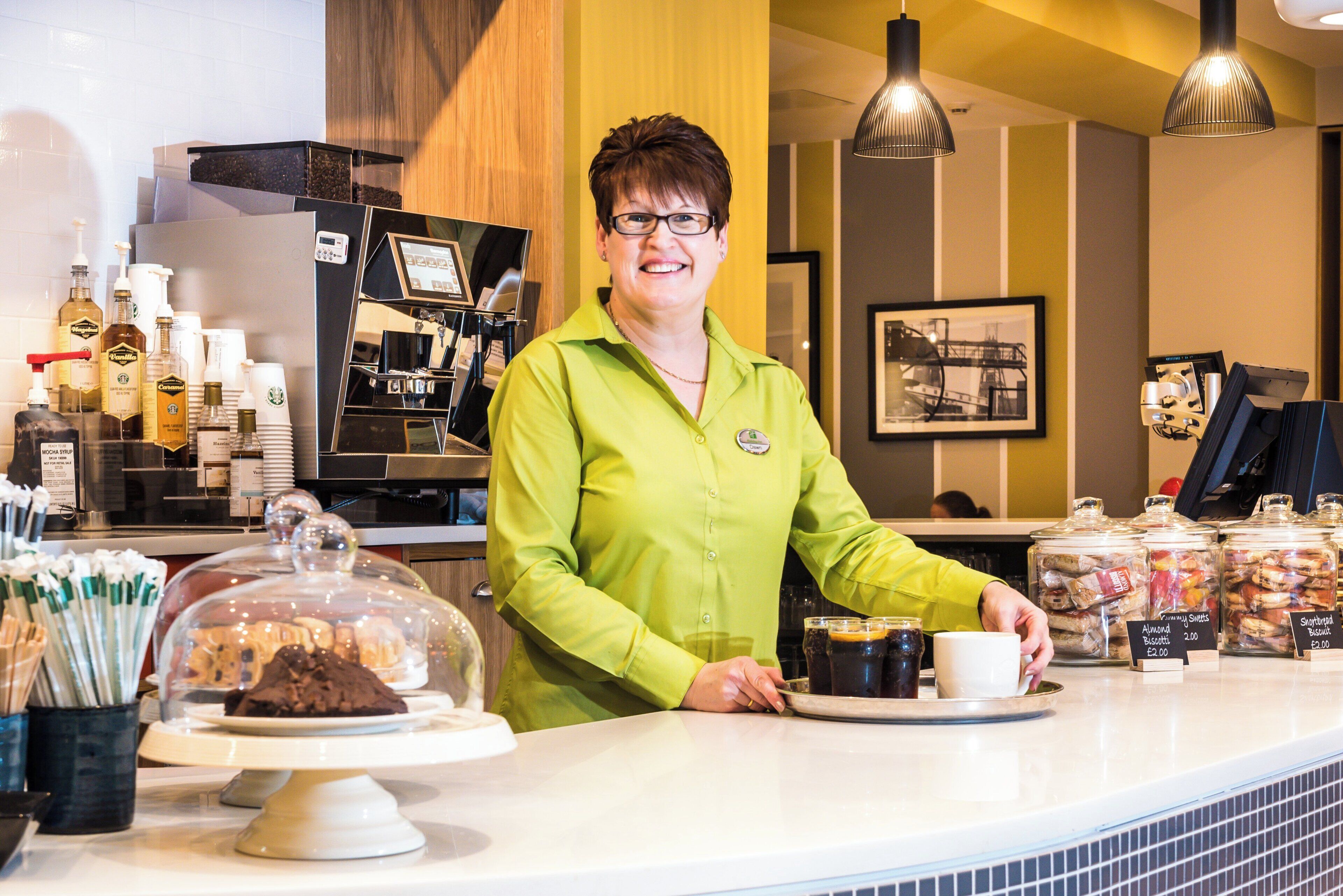A person standing behind a counter with a coffee machine, a display of pastries, and a coffee cup.