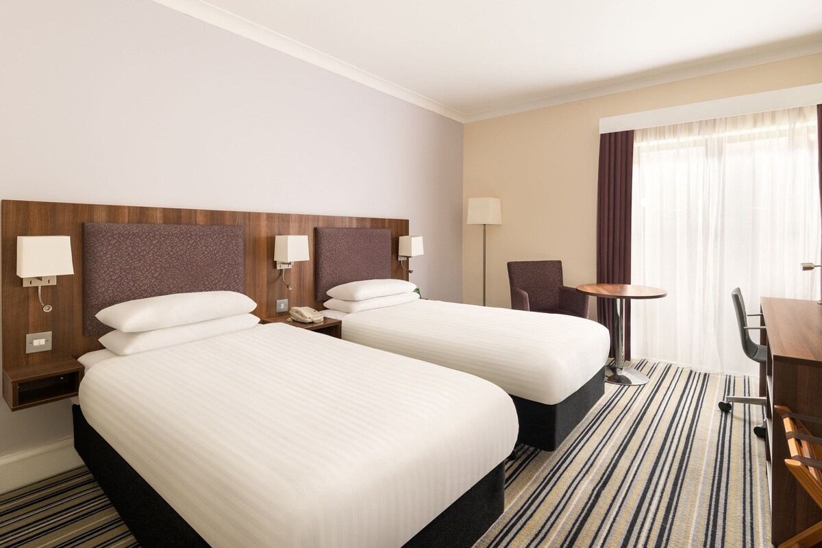 A hotel room with two beds, a desk, a chair, a TV, and a window with curtains.