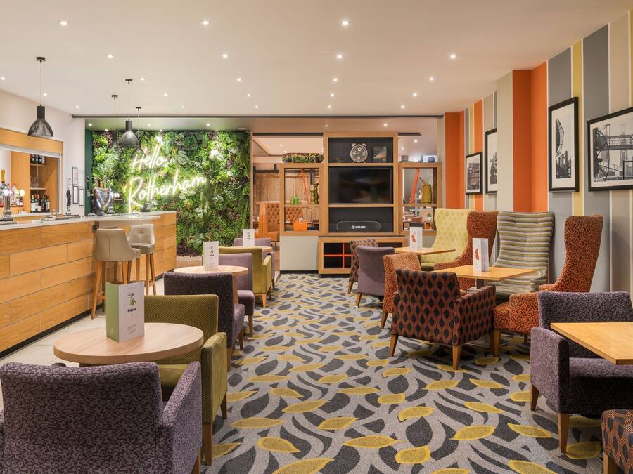 Un café moderne avec un comptoir en bois, plusieurs espaces de détente dotés de chaises à motifs et un mur végétal agrémenté d'un néon.