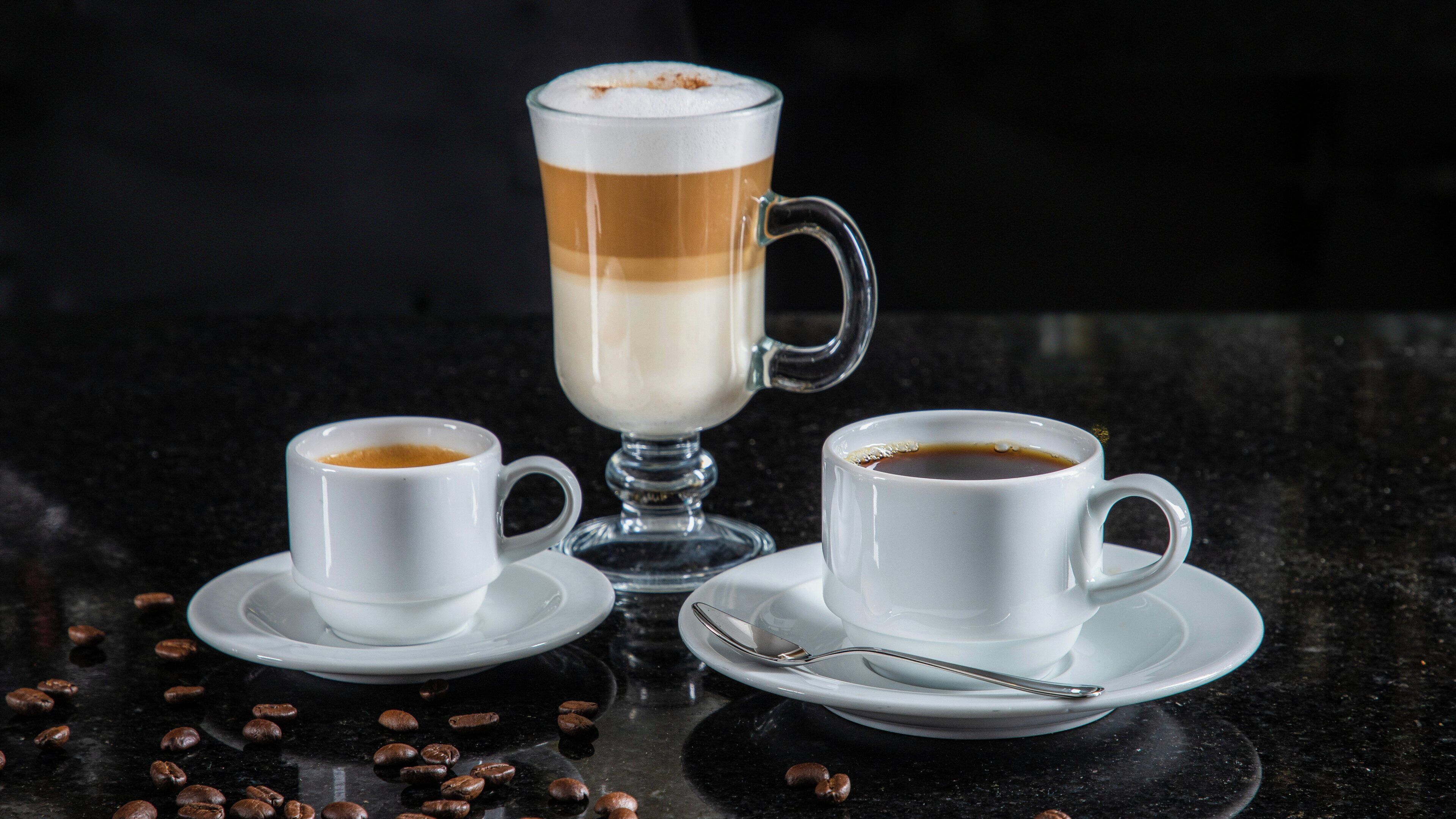 Three white cups of coffee on a black surface with coffee beans scattered around.