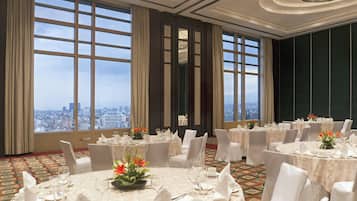 Un salón de banquetes con mesas redondas, sillas blancas y grandes ventanales con vistas a la ciudad.