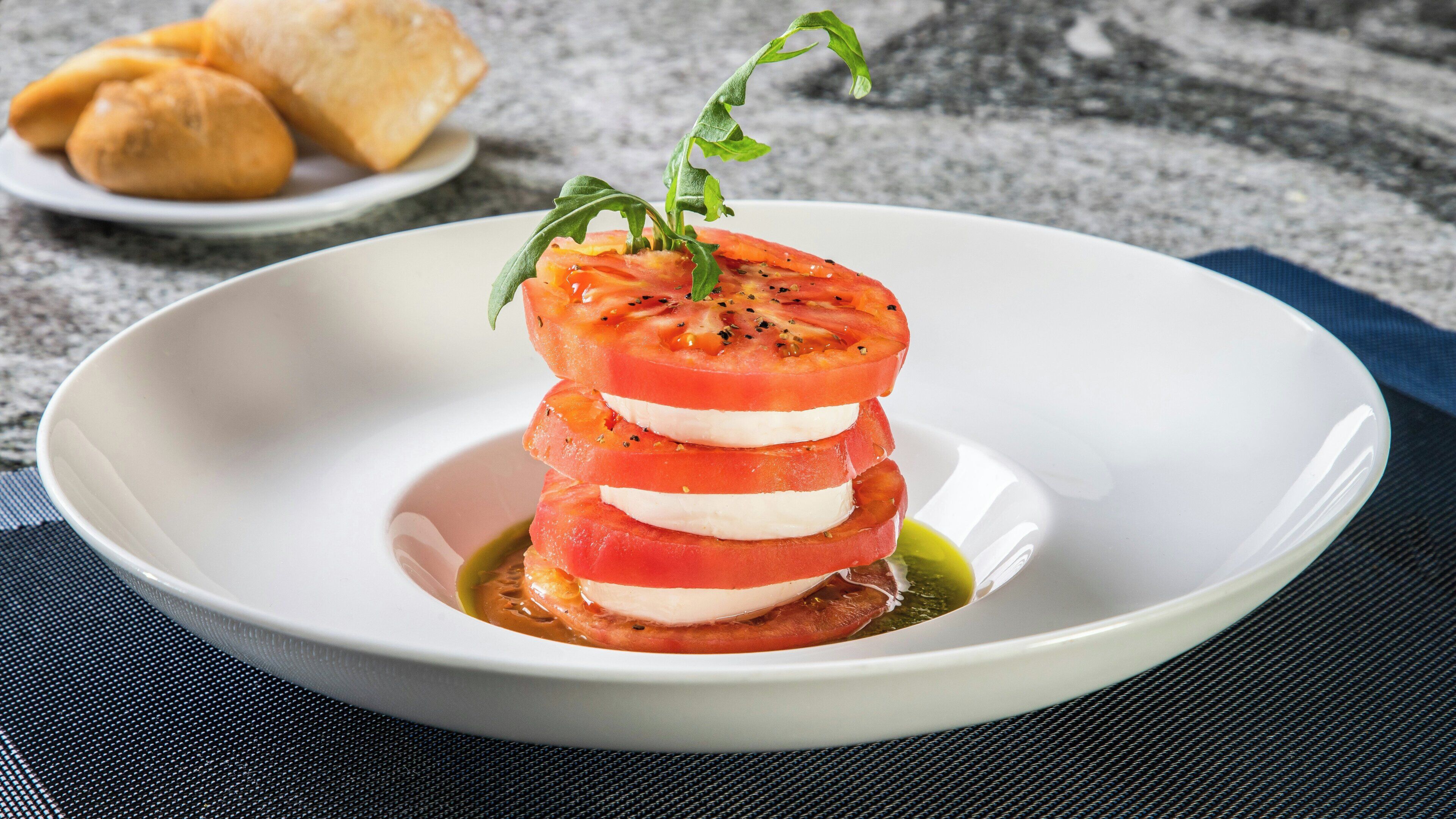Ein weißer Teller mit einer Schichtanordnung aus Tomaten und Mozzarella, garniert mit Basilikum, auf einer strukturierten Oberfläche.