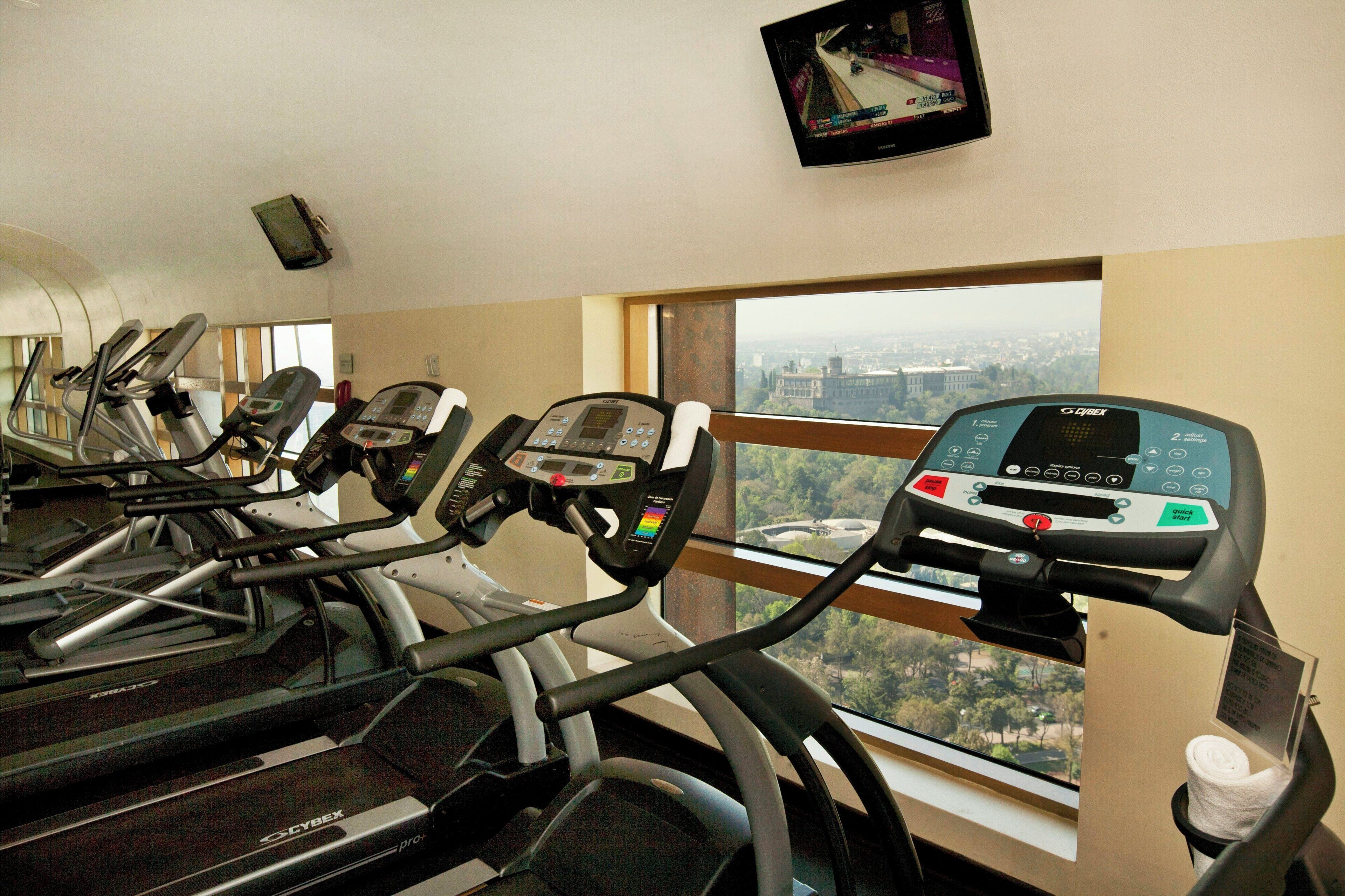 A gym with treadmills, a TV screen, and a view of a cityscape through large windows.