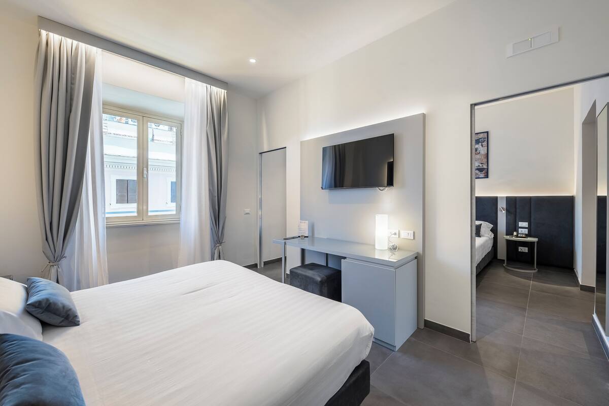 A modern hotel room with a large bed, a desk with a flat-screen TV, a small seating area, and a separate sleeping area with a bed.